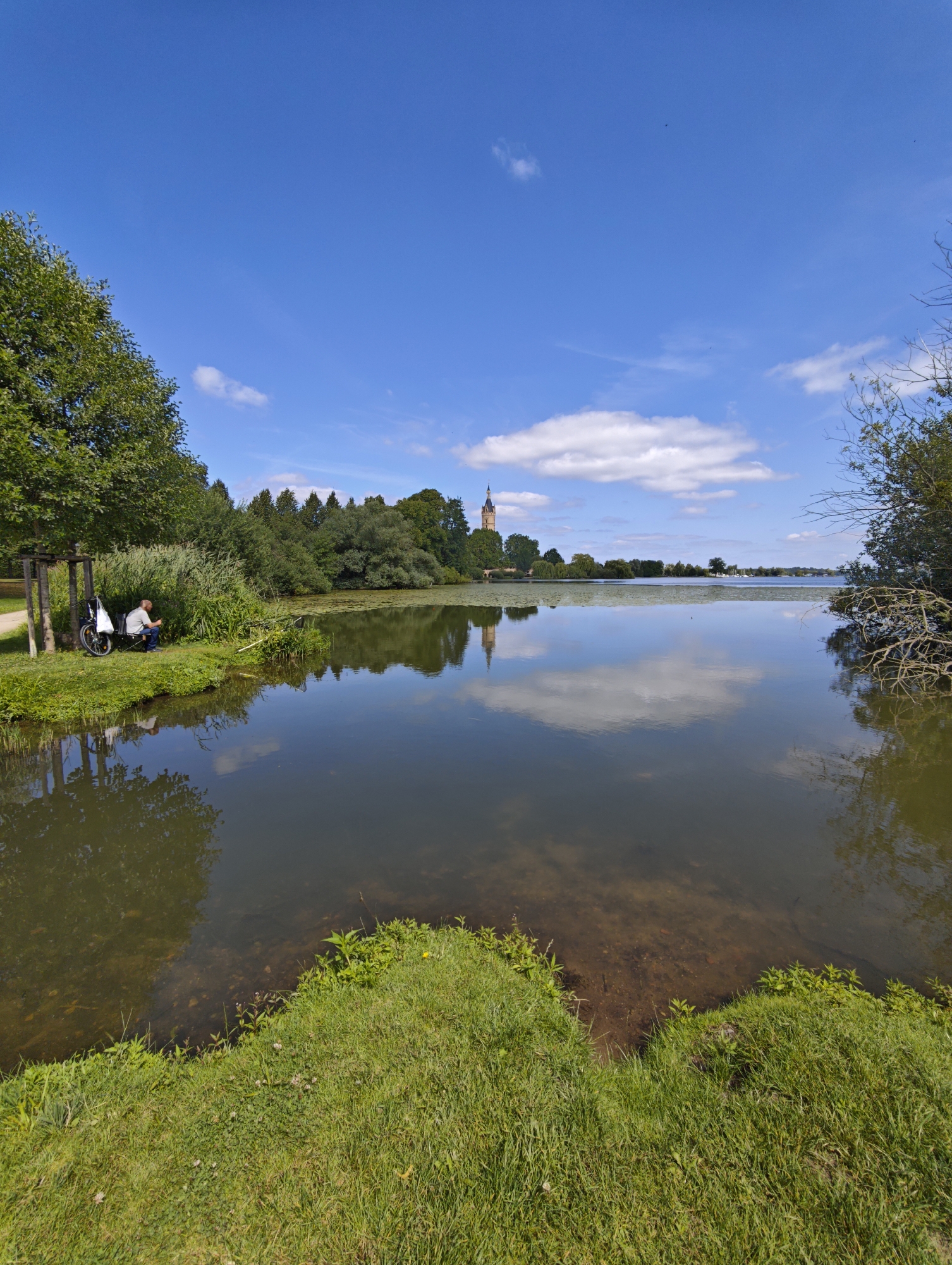 This is a wide-angle, eye-level photograph of a calm lake on a sunny day, likely located in the city of Schwerin, Germany, based on the presence of a distant tower that resembles the Schwerin Castle tower. In the foreground, lush green grass lines the edge of the water. To the left, a person is seated on a folding chair next to a bicycle with a white bag attached. This individual appears to be fishing, as a fishing rod is extended over the water. The lake is placid, reflecting the bright blue sky, scattered white clouds, and the surrounding trees and vegetation. In the middle ground, a line of dense green trees separates the lake from a faint outline of buildings and a prominent tower in the distance. The water is clear enough in places to see the sandy bottom near the shore.