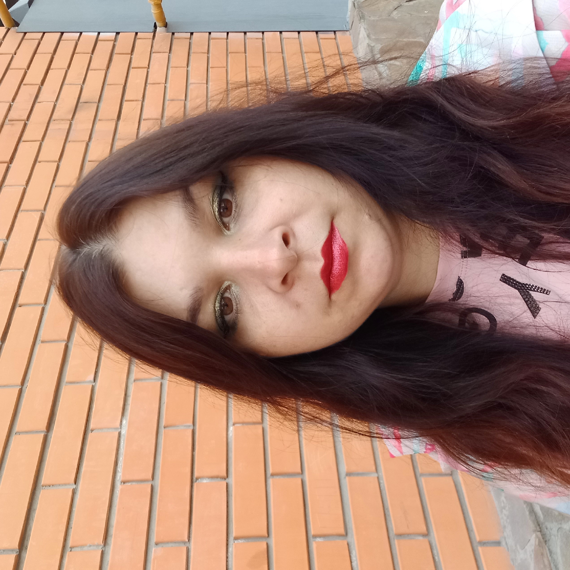 A medium shot image shows a woman with long, dark brown hair and light brown eyes posing in front of a brick wall. She is wearing makeup, including eyeshadow and red lipstick. She is also wearing a pink t-shirt with some sort of black graphic design on it. The background is an orange brick wall with grey mortar. To the right of the woman, a sliver of a grey metal fence is visible. The scene appears to be outdoors, possibly in a city. The location is Kropyvnytskyi, Ukraine.