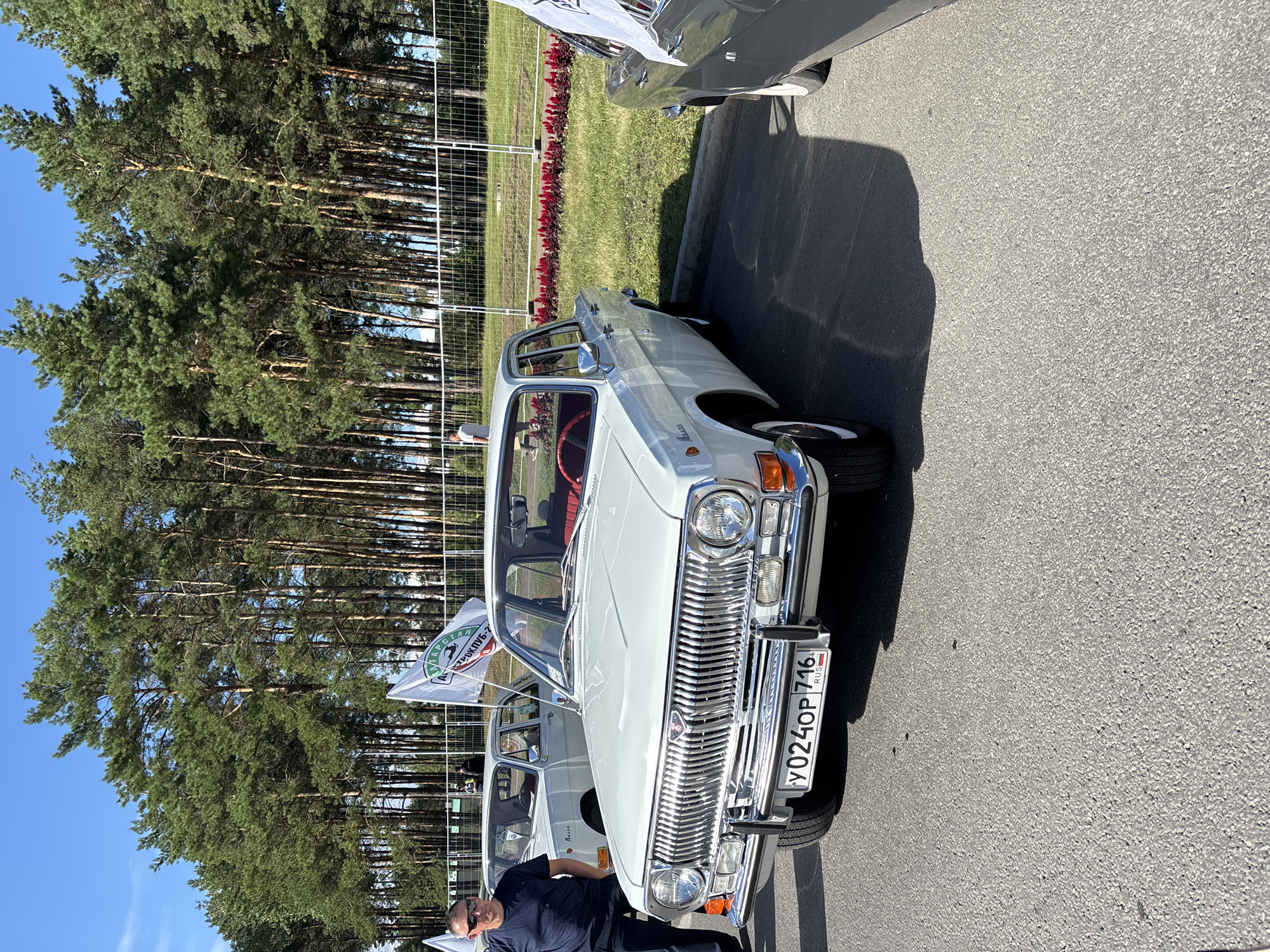 The image captures an outdoor car show on a sunny day, possibly in Naberezhnye Chelny, Russia, given the context. The primary subject is a white vintage Volga sedan parked on asphalt. The car features a prominent chrome grille, round headlights, and a red interior visible through the windshield. A license plate reading "Y024OP 716" is affixed to the front. To the left, a man wearing sunglasses, a dark blue t-shirt, and dark pants stands next to the car. Another white vintage car is visible behind it. In the background, a line of tall pine trees forms a backdrop, with a white metal fence separating the cars from a grassy area with red flowers. A white flag with a circular logo is mounted on the car in the foreground. Shadows suggest the sun is high in the sky.
