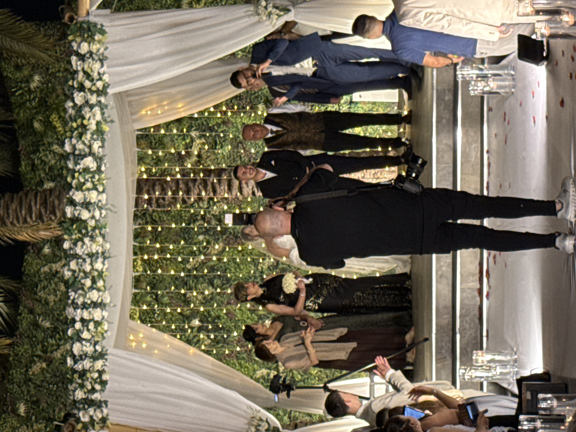 A wedding ceremony is taking place outdoors, likely in the Beer Tuvia Regional Council, Israel, at night. A chuppah adorned with white flowers and lush greenery frames the scene, with strings of warm-toned lights creating a festive backdrop. The bridal party stands on a raised platform draped with white fabric, while guests are seated on either side of a white-carpeted aisle. A photographer in black attire is positioned in the foreground, capturing the event. The groom and other male participants are dressed in suits, while the women wear elegant dresses. The atmosphere suggests a joyous celebration.