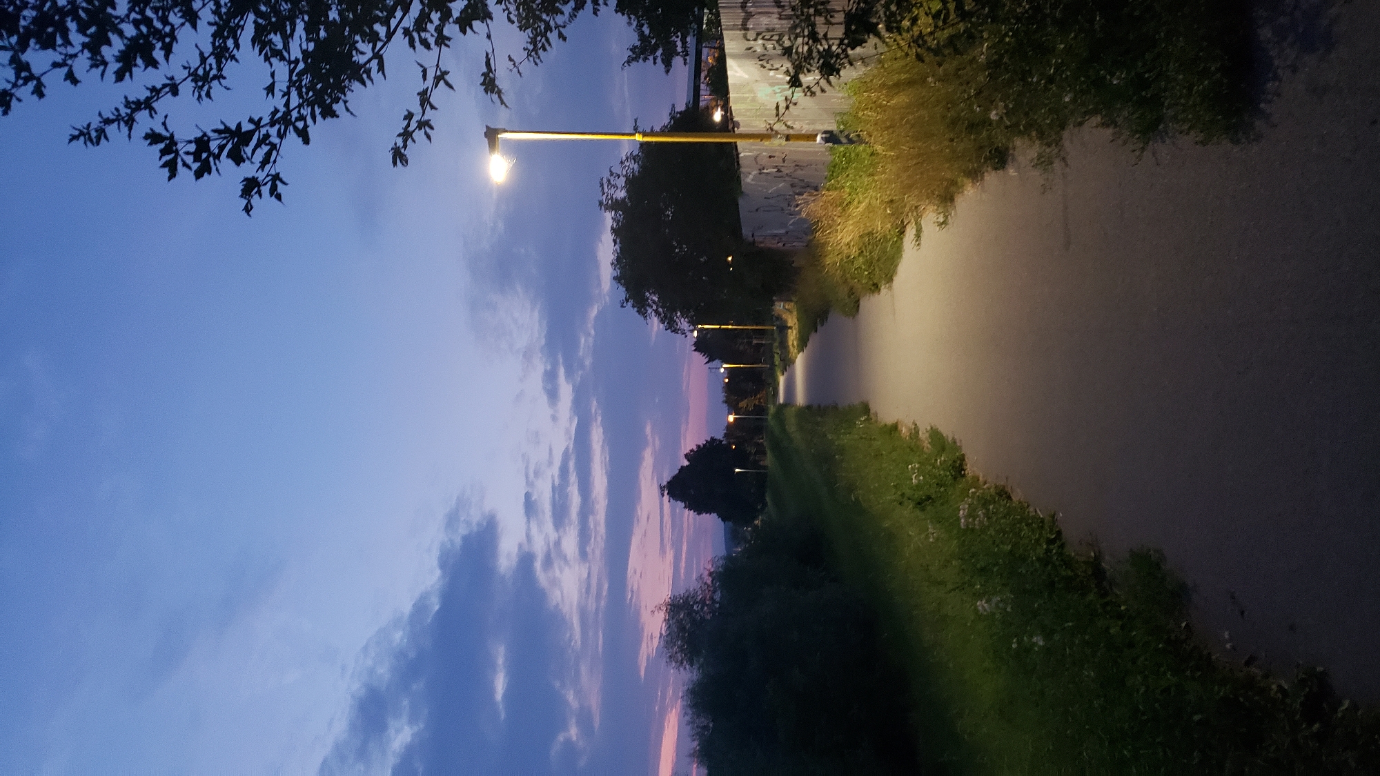 The image depicts a paved pathway lined with grass and vegetation, illuminated by streetlights. The sky above is a gradient of twilight colors, transitioning from deep blue to purple and pink hues, with scattered clouds. The pathway appears to be a pedestrian or cycling route, possibly in a park or recreational area within the city of Prešov, Slovakia, given the context provided. On the right side of the pathway, there is a fence with graffiti and tall grass. The overall scene suggests an evening or dusk setting, with artificial lighting providing visibility along the path. There are no people or animals visible in the image.