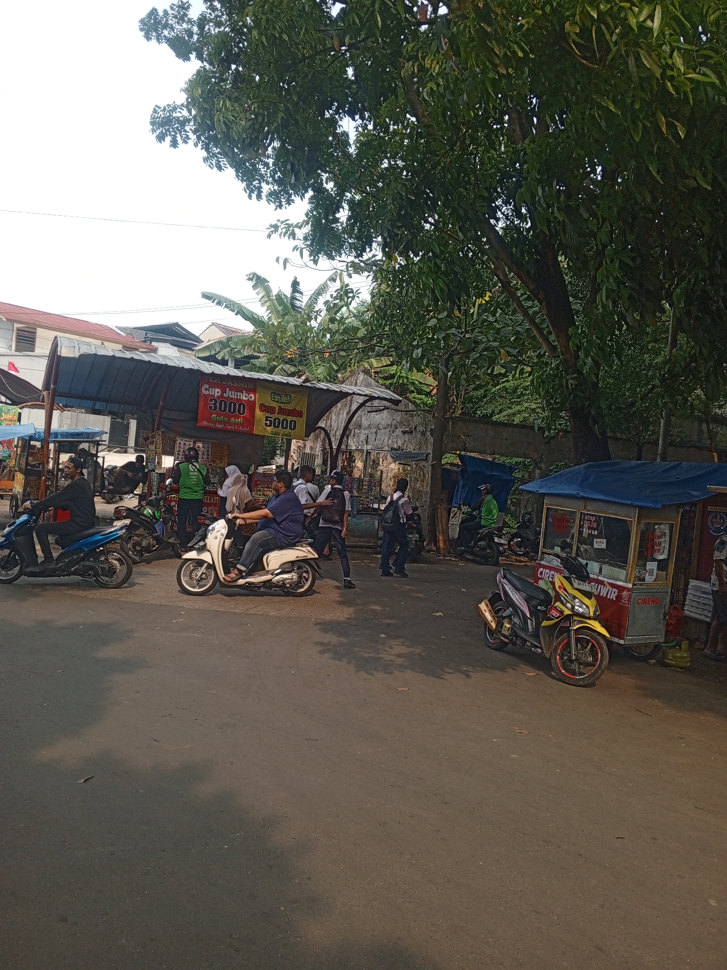 This is an eye-level, medium shot of a street scene in Depok, Indonesia, likely in a market or roadside stall area. The foreground features an asphalt road with visible shadows from trees and possibly some scattered debris. In the midground, several motorbikes are parked and in motion, indicating a busy area. One man in a dark shirt is riding a blue motorbike. Another man in a blue shirt is on a white scooter. A yellow motorbike is parked near a food stall. The food stall has a red and white body with a blue canopy, displaying signs with text in Indonesian, including "CIRENG" and "GULAI." Behind the stall, more people are visible, some interacting, and others appearing to be customers or vendors. Further back, there are more makeshift stalls or covered areas with awnings, and buildings with red roofs. The background is dominated by lush green trees and a bright sky, suggesting it is daytime. The overall atmosphere appears to be a typical urban street scene in Indonesia, with a focus on local commerce and transportation.