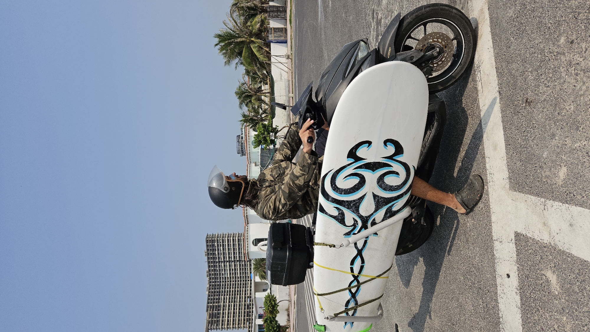 The image shows a person on a black motorcycle, transporting a surfboard, in Bac Cam Ranh, Vietnam. The person, seen from the left side, wears a black helmet with a clear visor, a camouflage-patterned jacket, and dark shorts, with black sandals on their feet. They are seated on the motorcycle, gripping the handlebars.
A white surfboard with a distinctive black and blue tribal-style design is attached along the right side of the motorcycle, secured by yellow and blue straps. A black storage box is mounted on the rear of the motorcycle.
The setting is an outdoor paved area, likely a road or parking lot, with white lines on the asphalt. In the background, there are several palm trees and multi-story buildings, some appearing partially constructed or unfinished, under a clear, bright blue sky. The strong shadows cast by the subjects indicate a sunny day, likely mid-morning or afternoon. There is no visible text in the image.