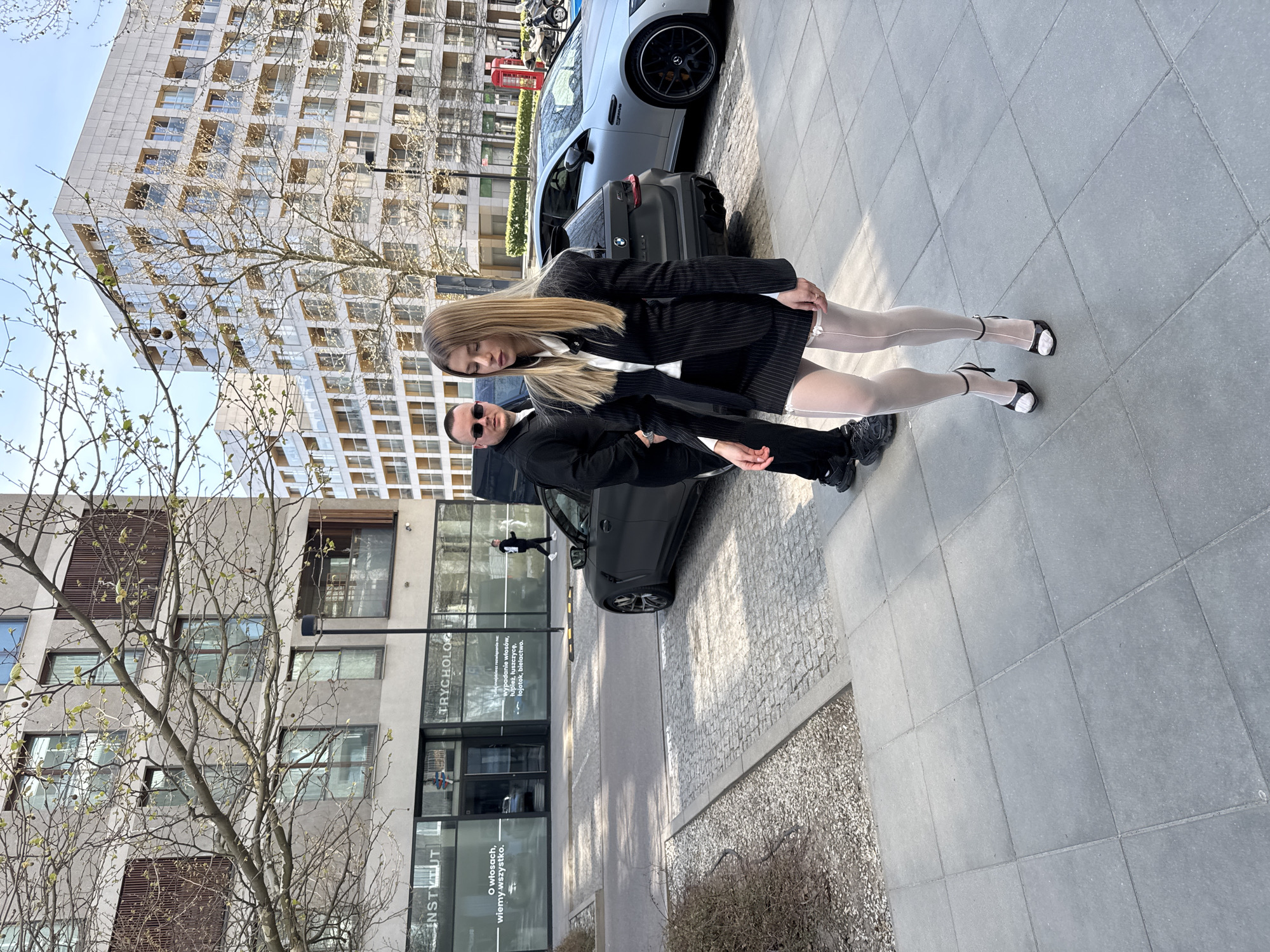 A man and a woman are standing in front of a car on a city street. The woman has long blonde hair and is wearing a black pinstripe suit jacket and a white shirt. Her legs are covered with sheer white hosiery and black Mary Jane shoes. The man is wearing a black suit and sunglasses. There is a gray car behind them and a building in the background. The storefront of "Instytut Tricholog," located in Warsaw, Poland, is visible behind the man. The storefront advertises services related to hair care.