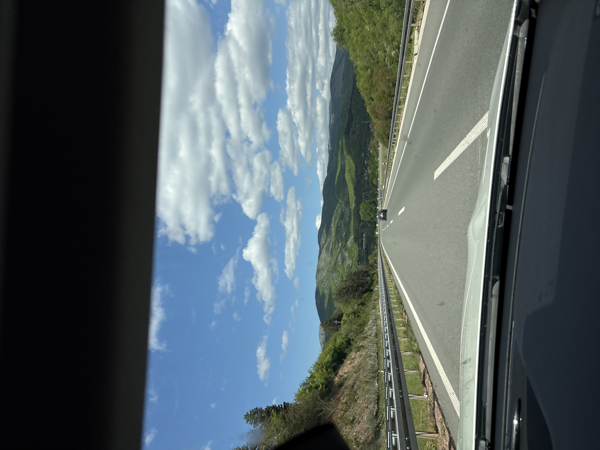 A highway stretches into the distance, framed by the interior of a vehicle. The road is marked with white lines and bordered by metal guardrails. Lush green mountains rise on the left side of the road, contrasting with a bright blue sky filled with fluffy white clouds. Another car is visible far ahead on the highway. The image was taken in Bela Palanka Municipality, Serbia.