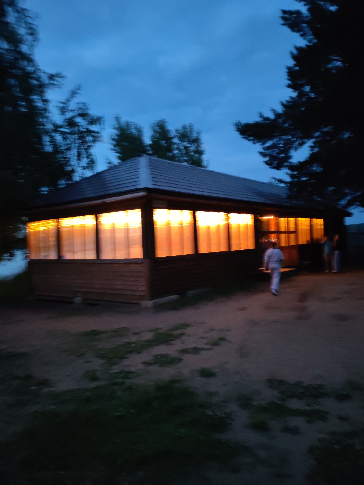 An exterior shot taken at dusk shows a wooden gazeboo with large windows, illuminated by a warm, orange glow from within. The gazeboo has a dark, sloped roof and is situated on a sandy, somewhat overgrown patch of ground. Three people are visible outside the gazeboo. One person, wearing a light-colored outfit that appears to be a martial arts uniform, walks away from the camera towards the entrance of the gazeboo. Two other figures, one in a blue top and the other in a dark top, stand near the entrance. Tall, dark trees frame the scene on the left and right, and the sky is a deep blue with some clouds. The overall impression is of a tranquil, late-evening gathering.