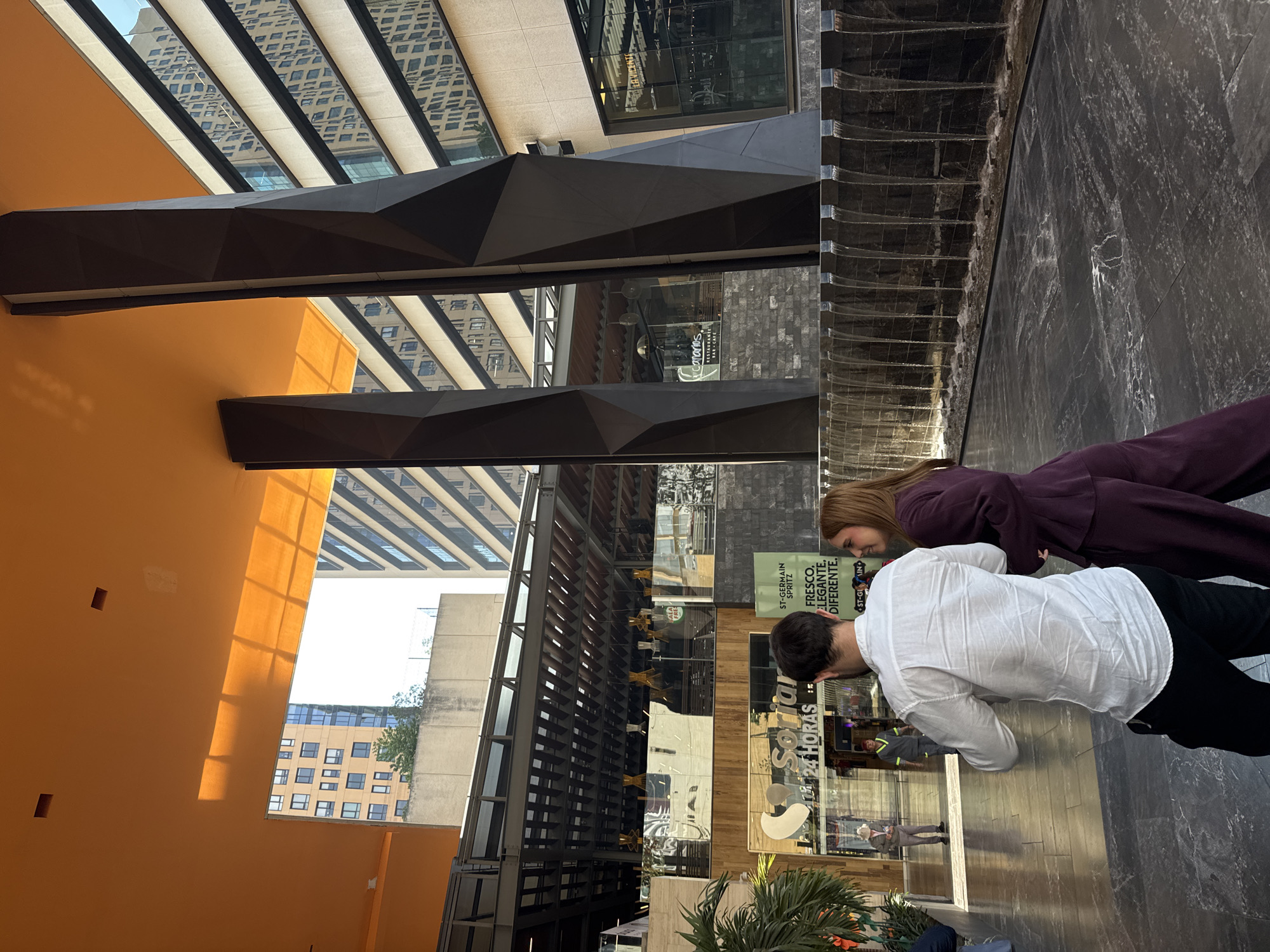 This media file captures a contemporary urban public space, likely a shopping center or commercial complex in Mexico City, Mexico. In the foreground, a man in a white shirt and black pants, and a woman in a deep purple outfit, are leaning forward and looking downwards, seemingly observing something on a lower level or a display.
The setting is characterized by striking modern architecture. A prominent, smooth orange wall dominates the left, with geometric shadows cast upon it by unseen structures. Large, black, angular sculptural elements, possibly parts of walkways or decorative beams, traverse the space. The ceiling features parallel light-colored beams with dark lines, reflecting a checkered pattern from an overhead structure. To the right, a dark marble wall features a cascading water element, creating a visually interesting texture. Beyond the figures, large glass storefronts reflect the surroundings and reveal glimpses of other people and commercial interiors. Tall, multi-story buildings with various window patterns are visible through and reflected in the glass.
Visible text includes:
* "Soniar 24 HORAS" on a storefront.
* An advertisement for "ST-GERMAIN SPRITZ FRESCO, ELEGANTE, DIFERENTE." with "ST-GLIN" also visible.
* "canarios RESTAURANTE BAR CAFE" on another storefront.
* "LA VICENTE" and "ALA" are partially visible on other signs.
The natural light and sharp shadows suggest it is daytime, with bright conditions outside. The overall atmosphere is modern, bustling, and sophisticated, indicative of a vibrant urban environment.