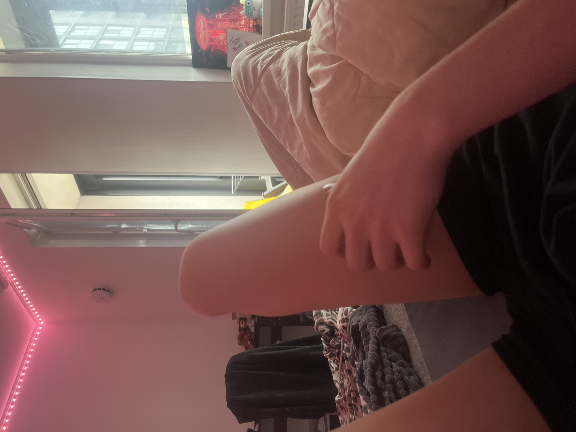 A person is seated on a bed in a room, with a window visible in the background. The room has a pink wall with LED lights and a smoke detector on the ceiling. Other objects in the room include a blanket with a distinct pattern and a black bag/clothing item. There is no indication of the location being Saint Petersburg, Russia.