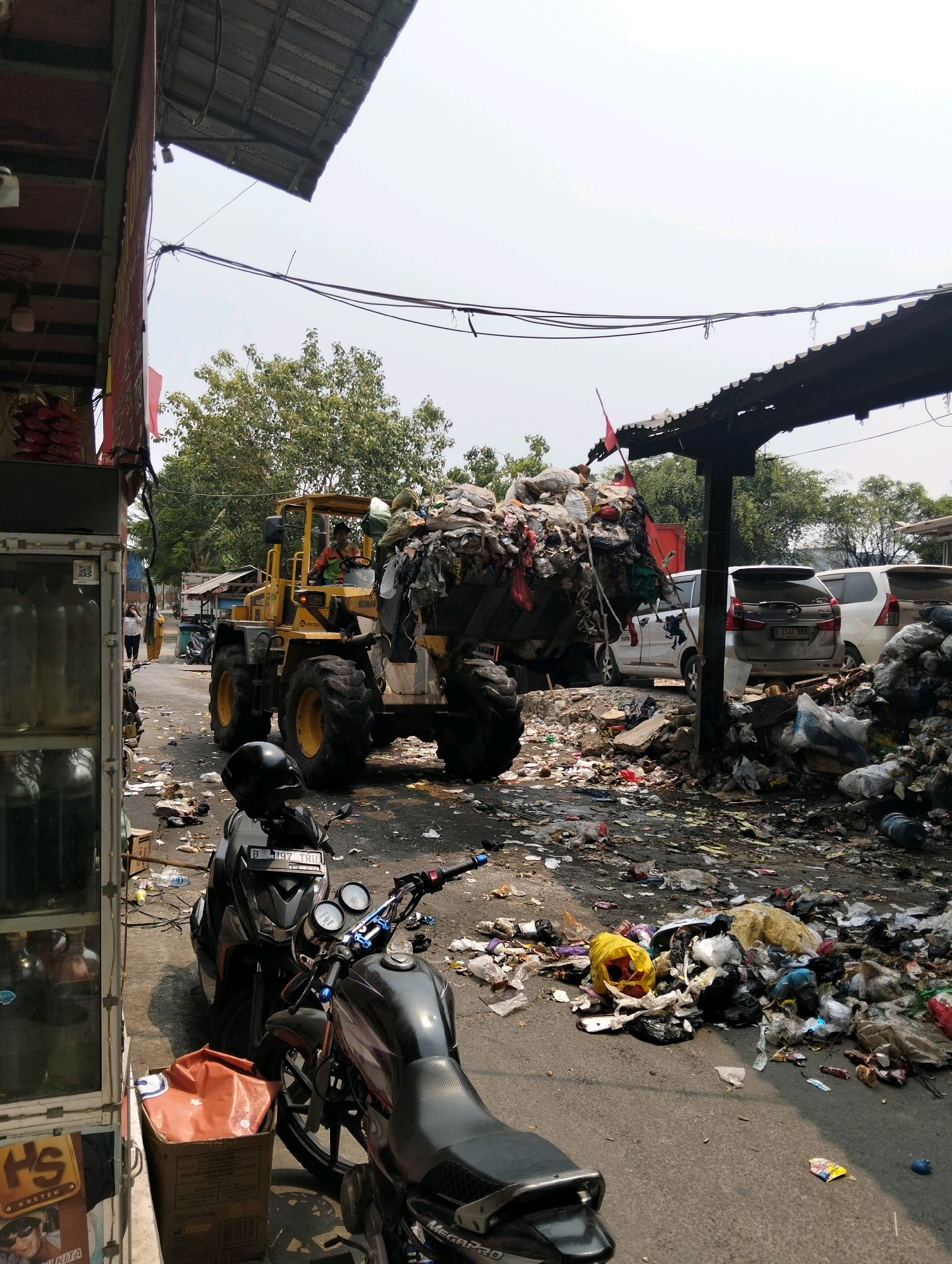 A yellow bulldozer is in the process of dumping a large pile of garbage onto an already debris-strewn street. The bulldozer is loaded with trash in its front bucket. The street is covered in loose garbage, including plastic bags, wrappers, and various other refuse. To the left of the bulldozer, a portion of a building with a glass display is visible, possibly a shop. In the foreground, two motorcycles are parked, one black and silver, and another partially visible black motorcycle. A cardboard box with an orange plastic bag inside is next to the motorcycles. In the background, several cars are parked, and trees can be seen behind them, suggesting an outdoor environment. The scene appears to be a waste disposal or processing area in an urban setting, likely in the city of West Jakarta, Indonesia, given the context. The sky is overcast.