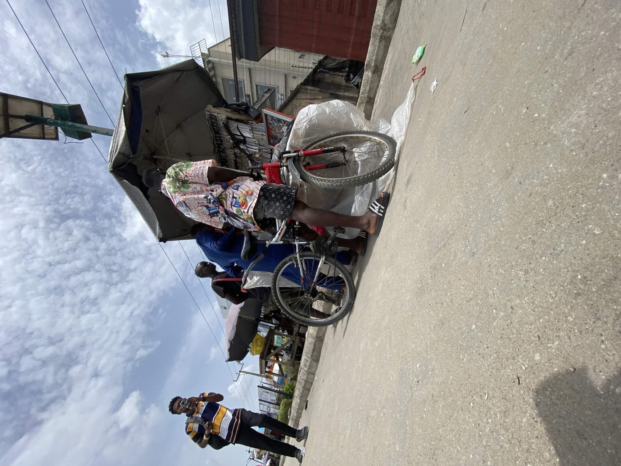 The image depicts a street scene in Lagos, Nigeria. The foreground features a man in a striped shirt holding a phone, likely taking a picture. Behind him, a group of people are gathered around a bicycle repair stand, which is set up under a makeshift umbrella. The stand is crowded with various bike parts and accessories. In the background are buildings, power lines, and signs of urban activity. The street is paved, and the sky is partly cloudy.