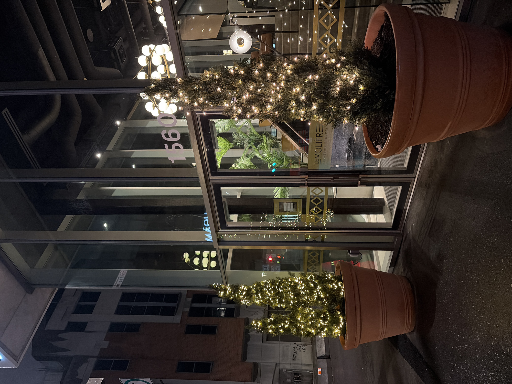 Here is a description of the image:
The image depicts the exterior of a business in Montreal, Canada. The business is called "Le Molière Brasserie Française," as indicated by the signage on the storefront. The building's facade is modern, featuring glass doors and windows. Large, terracotta-colored planters flank the entrance, each containing a small evergreen tree adorned with warm white string lights. The lights suggest it is either the holiday season or the establishment maintains a festive ambiance year-round. The building's address, "1560," is prominently displayed in large white numerals on the glass. The street outside appears to be wet, possibly from recent rain. The reflections in the glass show other buildings across the street. Overall, the scene conveys an upscale, inviting atmosphere.