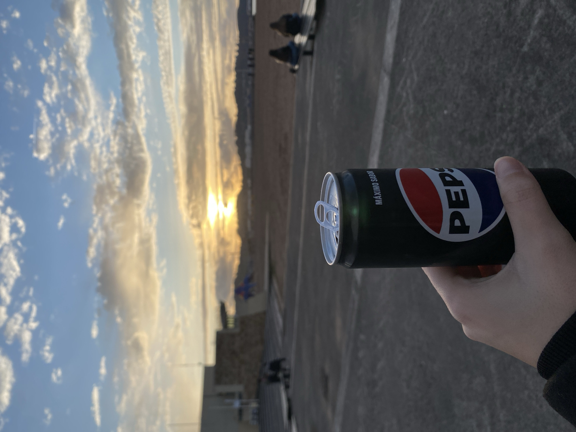 A person holds a black Pepsi can. The can has the words "MÁXIMO SABOR" written on it. The background shows a sky with a cloudy sunset and a beach. In the distance, blurred figures are sitting on benches. The location appears to be near San Lorenzo Beach in Gijón/Xixón, Spain, based on the terrain and presence of beach-related features. The person is holding the can in their right hand. The can is unopened. The sky suggests it may be late afternoon or early evening. The beach is visible behind the person, and blurred individuals are sitting on benches in the vicinity. The ground appears to be paved.