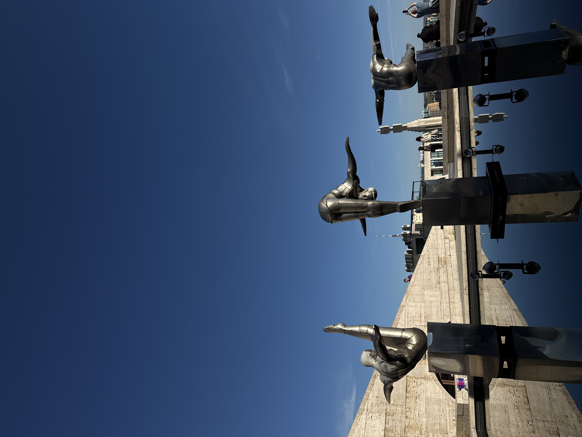 The image depicts the Cascade complex in Yerevan, Armenia. The foreground features three metallic sculptures of human figures in dynamic poses, seemingly suspended in mid-air. The sculptures are mounted on dark, rectangular bases. The backdrop is a clear, deep blue sky. In the background, partially visible, is a view of Yerevan's skyline, including buildings and structures. Pedestrians are seen in the distance. The photograph is taken from a low angle, emphasizing the height and scale of the Cascade complex and its sculptures. The overall scene is a sunny, urban environment with a focus on the architectural and artistic elements of the city.
