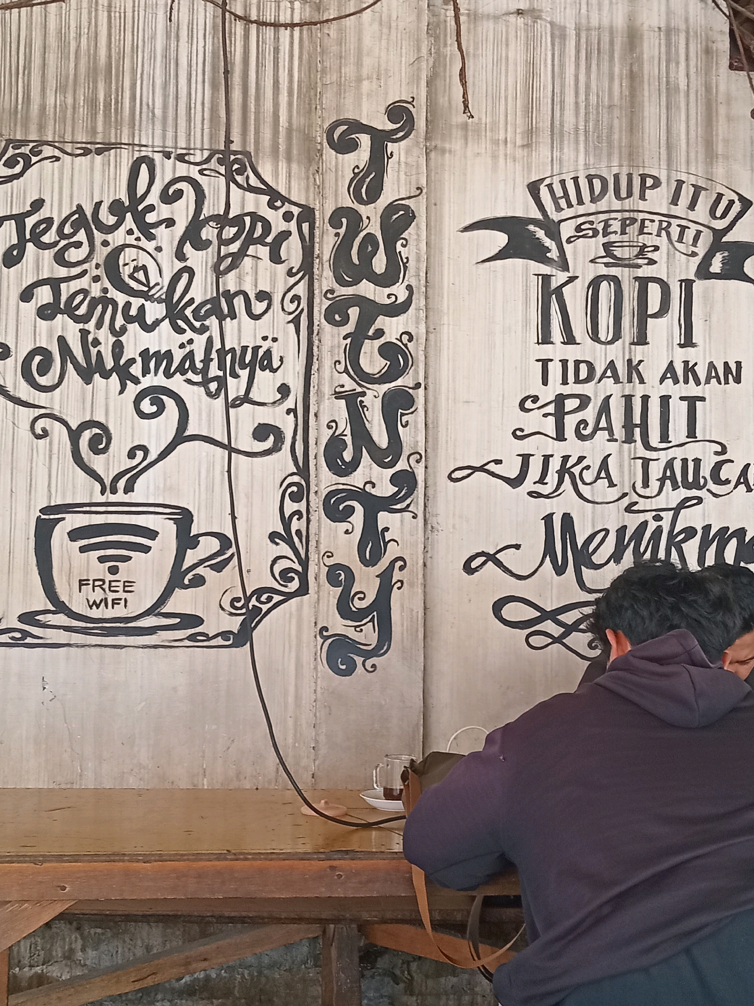 In Ngipik, Indonesia, a wall in a cafe is adorned with large, black, handwritten calligraphy. To the left, the words "Jelajahi Kopi," "Temukan," and "Nikmati" are written, accompanied by an illustration of a coffee cup with steam rising and the words "FREE WIFI." To the right, a banner reads "HIDUP ITU SEPERTI KOPI," followed by "TIDAK AKAN PAHIT JIKA DITAU CARA MENIKMATINYA." Two individuals, their faces obscured, are seated at a wooden table in front of the wall, facing away from the camera. One is wearing a dark hooded sweatshirt. The cafe environment appears rustic with visible wooden textures.