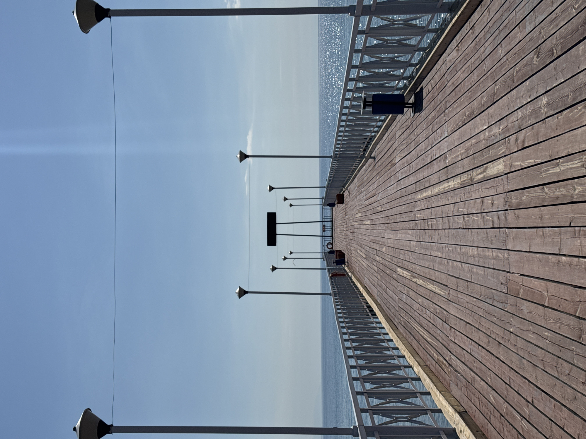 A long wooden pier extends into a calm, blue body of water under a clear sky. The pier is lined with a metal railing featuring an ornate diamond pattern. Several lampposts are spaced along the railing. At the end of the pier, a dark rectangular sign is visible, supported by green posts, with a life preserver hanging on its side. A blue trash receptacle is located on the right side of the pier. The water reflects the bright sunlight, creating a shimmering effect. The scene appears to be located in the city of Koshkol', Kyrgyzstan, likely a public recreational area by the water. There are no people visible in the image.