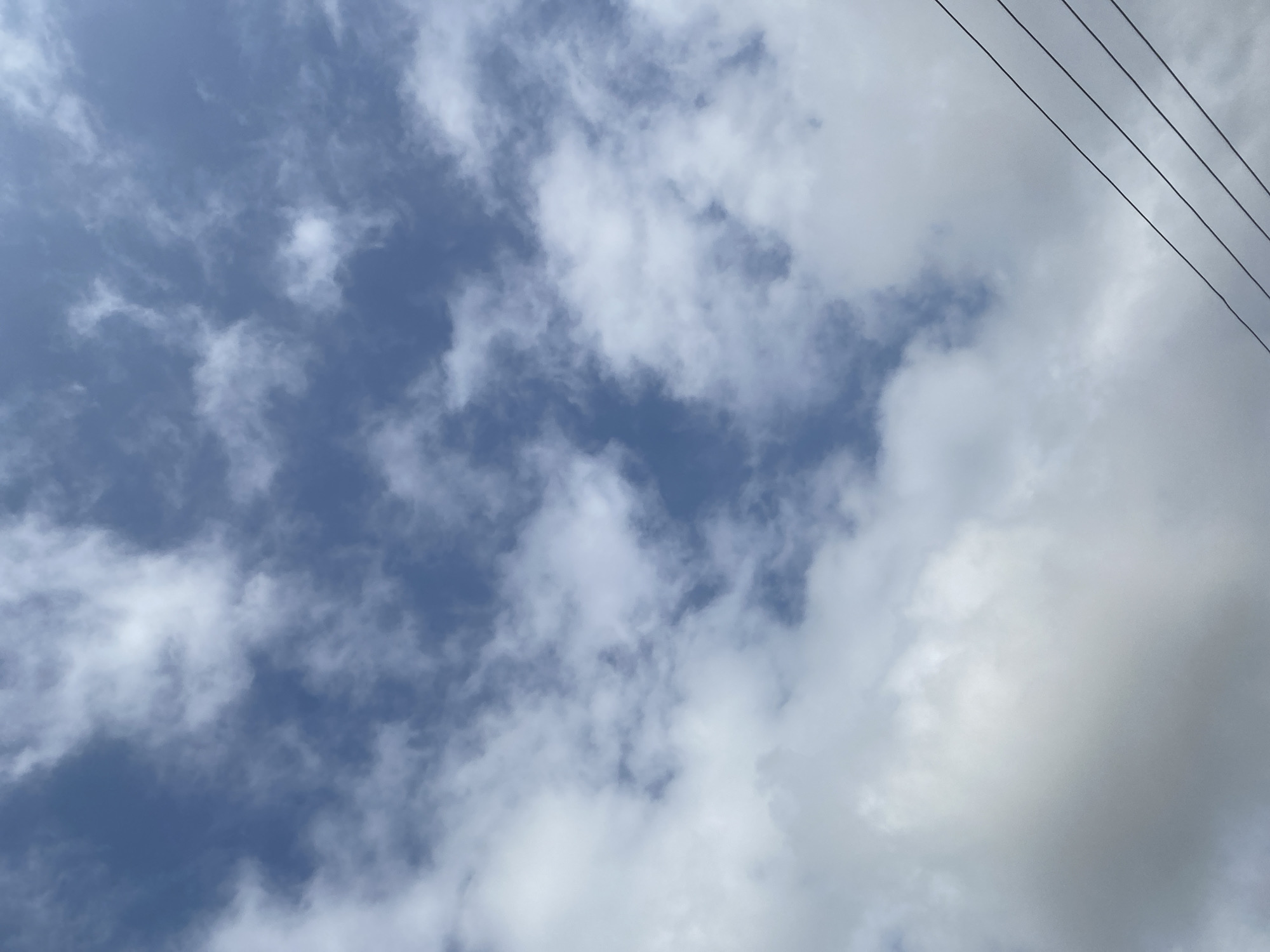 The image shows a view of a cloudy sky. The clouds are a mix of white and gray, with patches of blue sky visible between them. In the lower right corner of the image, there are four parallel black wires stretching diagonally across the frame, suggesting the presence of power lines. The scene appears to be an outdoor setting, likely in Lafia, Nigeria, given the context of the prompt. There are no people or specific objects or actions depicted, beyond the natural elements of the sky and the man-made power lines.