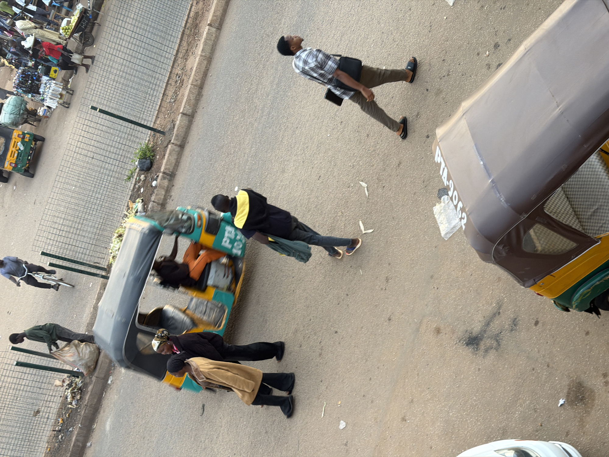 This is a high-angle, medium shot of a street scene in Bukuru, Nigeria. In the foreground, a brown and green autorickshaw is parked. To the left of the autorickshaw, a person in a tan coat and black pants walks away from the camera. To the right of the autorickshaw, a man in a plaid shirt, khaki pants, and black slides walks to the left, carrying a black bag across his chest and a tablet in his right hand. In the middle ground, an autorickshaw is in motion, carrying two passengers. A person on a bicycle rides in the background, wearing a blue shirt and black pants. A man in a green shirt and gray pants is standing on the sidewalk, holding a large, light-colored plastic bag. The street is paved with asphalt and there is a metal fence bordering the sidewalk, which is littered with trash.