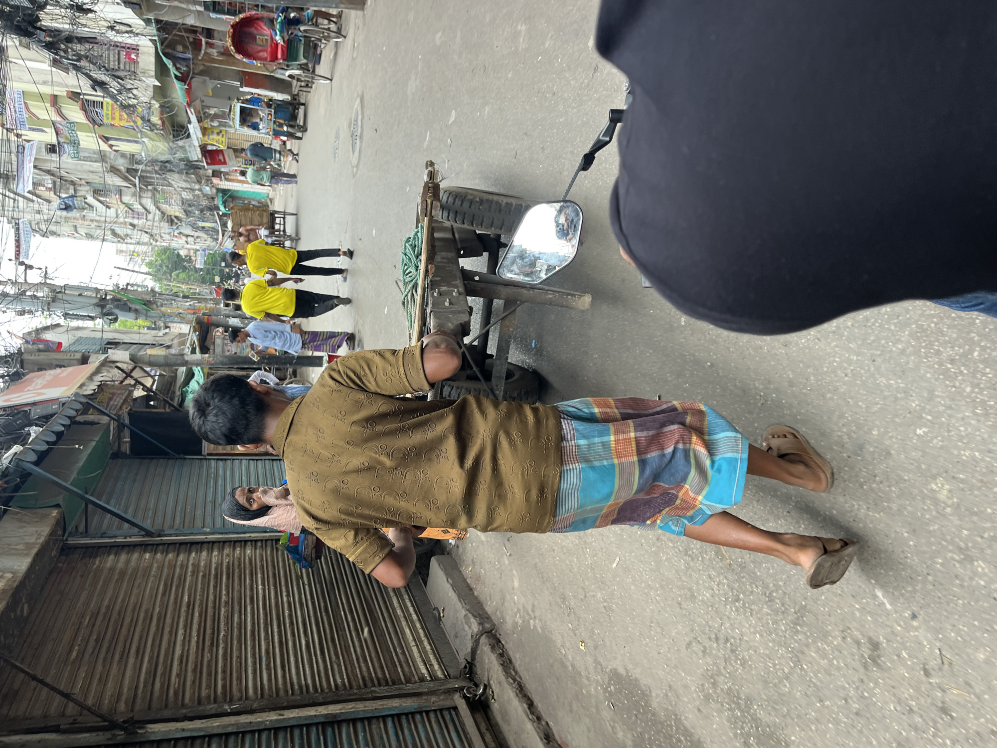 This image captures a street scene in Dhaka, Bangladesh. In the foreground, a person wearing a brown patterned shirt and a colorful lungi is pushing a two-wheeled cart laden with green rope. The cart has two large tires. To the left, a woman in a pink headscarf is visible. In the background, several people are walking along the street, including two men in bright yellow shirts and one in a light blue outfit. Buildings line both sides of the narrow street, with shops having their shutters down. Electrical wires are prominently strung overhead, characteristic of urban environments in Dhaka. The road surface appears to be asphalt, with some debris scattered on it. The overall impression is a busy, everyday street scene in a densely populated area.