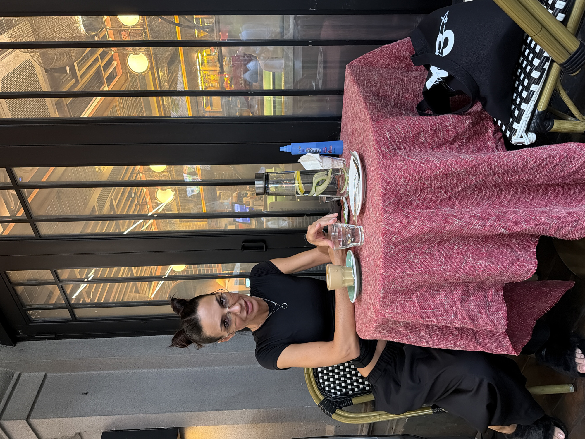 A woman sits at a small, round table outside a cafe, her legs crossed, wearing black. She has dark hair styled in two buns and is wearing glasses and a black t-shirt. On the table in front of her are a coffee cup, a glass of water, a carafe of water with cucumber and lemon slices, a small bottle of blue spray, and a plate with silverware. The table is covered with a pink and red textured tablecloth. A black tote bag with white lettering rests on the chair to her right. The background shows a large glass window with a black frame, reflecting the interior of the cafe which appears to be a restaurant with lights and suspended ceiling elements. The cafe is likely located in Chancheng District, China, given the context.