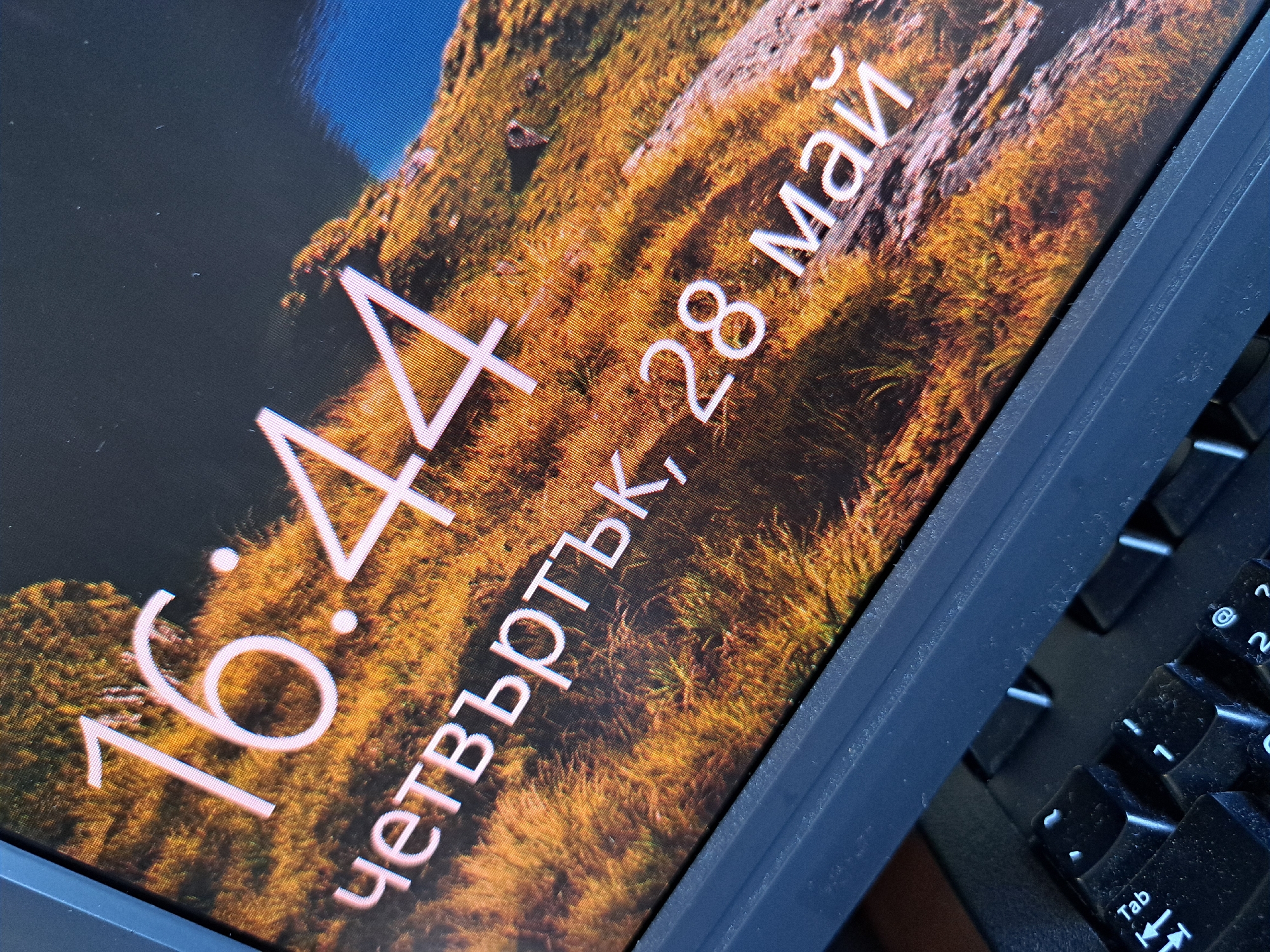 A close-up angled view shows a computer screen displaying the time as 16:44, the day of the week as "четвъртък" (Thursday), and the date as 28 май (May 28). The language appears to be Bulgarian. The background of the screen is a landscape image with a mix of colors, including browns, yellows, and blues. A black keyboard is partially visible to the right of the screen. Given the use of the Bulgarian language for the date and time, the image likely originates from Bulgaria, but the specific location within Sofia cannot be determined from the image alone.