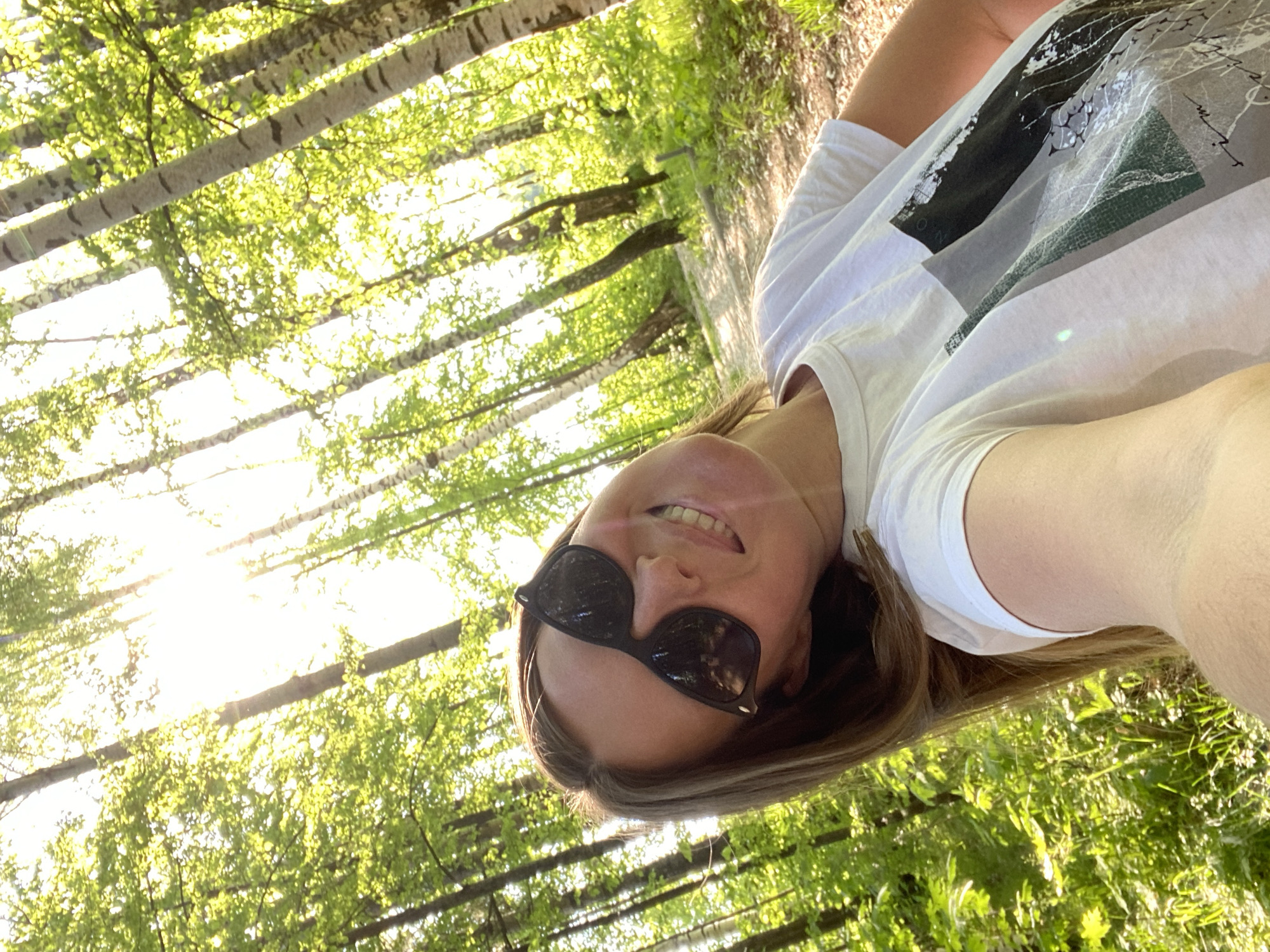 A fair-skinned woman with light brown hair is centered in the frame wearing sunglasses, partially visible with a white t-shirt. The background shows dense trees with green leaves and thin white trunks. The sunlight is shining through the trees. This photo was taken in a forest in Noviy, Russia.