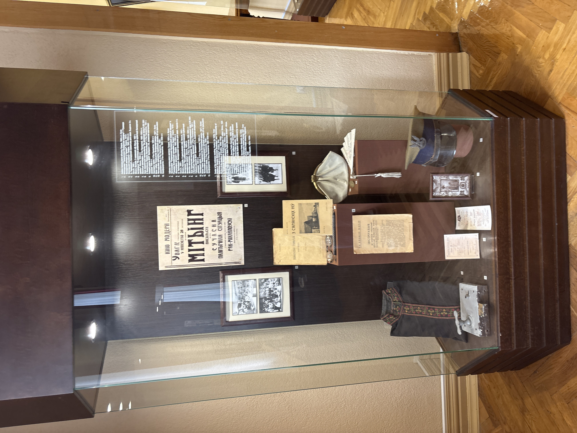 A glass display case containing historical artifacts is positioned in a museum setting in Hrodna, Belarus. The display case is made of wood and glass and is lit by spotlights from above. Inside the case, various items are arranged on shelves and stands. On the left side, there is a dark garment with embroidered trim, possibly a traditional coat or robe. Below it rests a metal object that appears to be a lock mechanism. To the right of the garment, there are several documents and a small framed picture. In the center of the display, a white satin clutch purse is placed next to a stack of what look like folded papers or fabric. Above the purse, there is a framed print or poster with text in Cyrillic and two black and white photographs. To the right of the purse, a military-style cap with an emblem is displayed on a stand. There are other assorted documents and smaller framed items scattered throughout the case. The background wall is a neutral beige color, and the floor is made of polished wooden parquet.