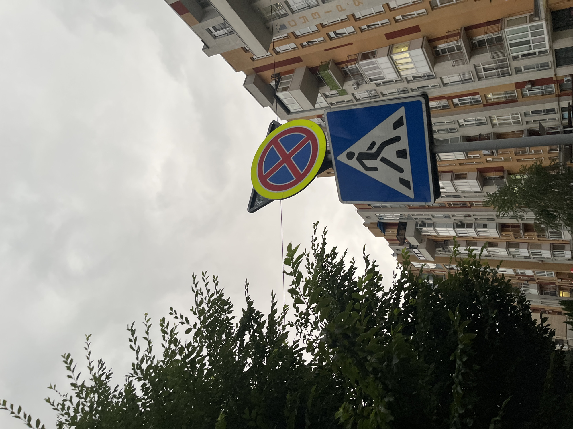The image depicts an outdoor urban scene with two prominent traffic signs. The upper sign, circular with a yellow border, shows a red prohibition symbol with a blue background and a red cross, indicating "no stopping." Below it, a triangular blue sign with a white border features a silhouette of a person walking on a pedestrian crossing, signifying a crosswalk. In the background, a multi-story residential building with a modern facade of orange, grey, and white elements and numerous balconies is visible. A large green tree with dense foliage occupies the left side of the frame, partially obscuring the sky, which is overcast with grey clouds. The text "СУЗІР'Я" is visible vertically on the building facade. The perspective is from a low angle, looking upwards towards the signs and the building. This scene is likely located in Chernivtsi, Ukraine, given the Ukrainian text on the building.
