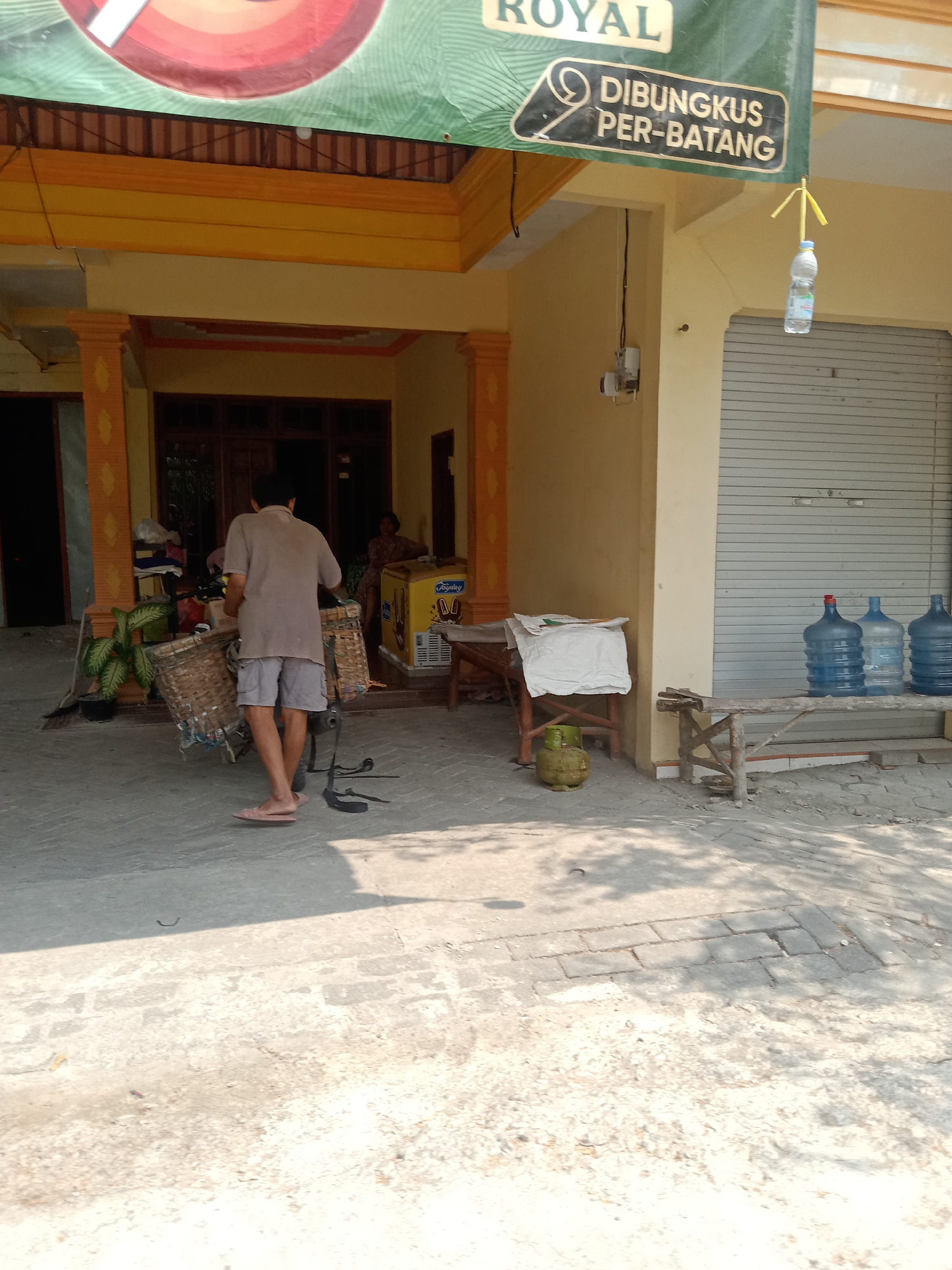 An exterior shot captures the front of a building with a yellow facade and an orange trim under a green awning that reads "ROYAL DIBUNGKUS PER-BATANG". A man in a light brown t-shirt and grey shorts is walking away from the camera, carrying a large woven basket. To his left, another woven basket is partially visible. In the background, a doorway leads into a building with glass doors. A person is seated inside. Next to the doorway, a white and yellow refrigerator with a "JAPRI" logo sits on the ground. To the right of the building's entrance, a long, rough wooden bench is set up against a closed, corrugated metal shutter. Three large blue water jugs are placed on the bench. In front of the bench, a small gas cylinder rests on the ground next to a wooden structure holding a white cloth. A single plastic bottle with a yellow straw is hanging from a string from the awning. The ground is paved with concrete and brickwork, with shadows cast by the overhead structures.