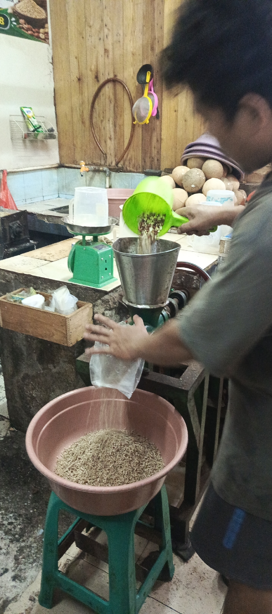 In a dimly lit room, a person's hands are visible, operating a grain processing machine in what appears to be a market or workshop in Sukaresmi, Indonesia. A person, partially visible from the shoulder up, is seen wearing a dark grey t-shirt and dark shorts. Their right hand is engaged in pouring grain from a green plastic funnel into a metal hopper attached to the processing machine. The left hand is holding a clear plastic bag, from which fine particles are falling into a large, light pink plastic bowl positioned on a green plastic stool. The bowl is already filled with a significant amount of processed grain.
The background features a wooden wall with various items hanging on it, including a brown circular object and a pink and yellow object that resembles a sieve or strainer. To the left, a green kitchen scale with a clear plastic container on top is visible, suggesting weighing operations. Several coconuts are piled up behind the person. A wooden box containing white plastic bags is also present on a counter to the left. The floor appears to be tiled and somewhat grimy, with some scattered debris. The overall environment suggests a functional, perhaps slightly unkempt, space dedicated to food processing.