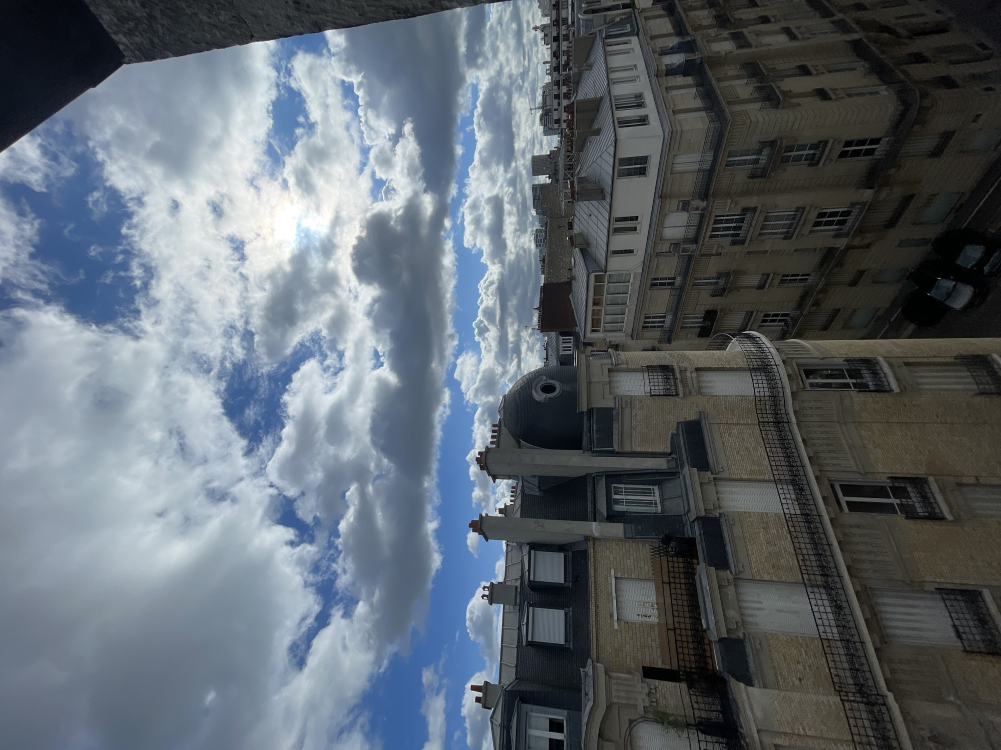 The image is an elevated, outdoor view of Parisian architecture under a partially cloudy sky. The perspective is from a high vantage point, looking down and across a street. In the foreground, the ornate mansard roofs and facades of Haussmannian buildings are visible, characterized by their stone construction, numerous windows with shutters, and decorative balconies. Chimneys are prominent on the rooftops. The sky above is a mix of bright blue patches and large, white, fluffy clouds, with the sun peeking through in the upper center of the frame, creating a bright, diffused light. In the distance, more contemporary buildings and construction cranes can be seen, indicating a mix of historical and modern urban development. The overall impression is of a daytime scene in a dense European city.