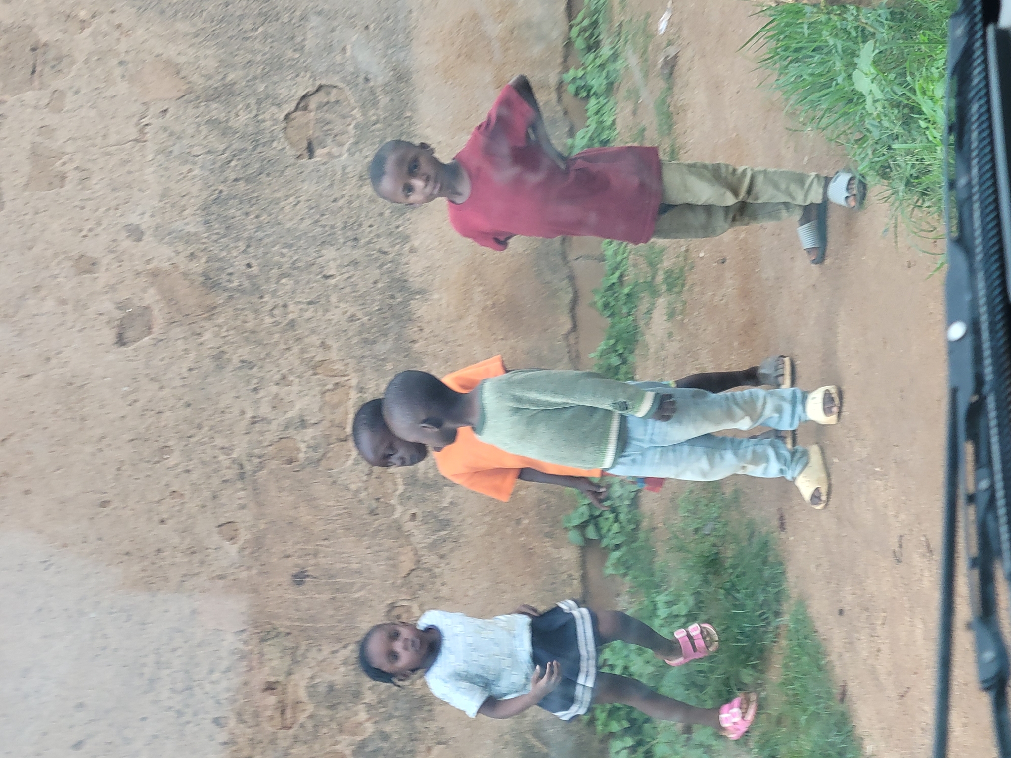 Four children stand on a dirt path in front of a rough, textured wall. A young girl on the left wears a light blue short-sleeved shirt and a dark pleated skirt. She has dark hair pulled back and pink sandals on her feet. Next to her, a boy in an orange t-shirt and blue jeans looks to his right. In the center, a boy in a light green long-sleeved shirt and blue jeans faces forward. To the right, a boy in a maroon t-shirt and light brown pants stands with his hands on his hips, looking towards the viewer. The environment suggests an outdoor setting, possibly in a less developed area, consistent with parts of Jos, Nigeria, where unpaved surfaces and rustic building materials are common. The lighting is even, indicating it is daytime.