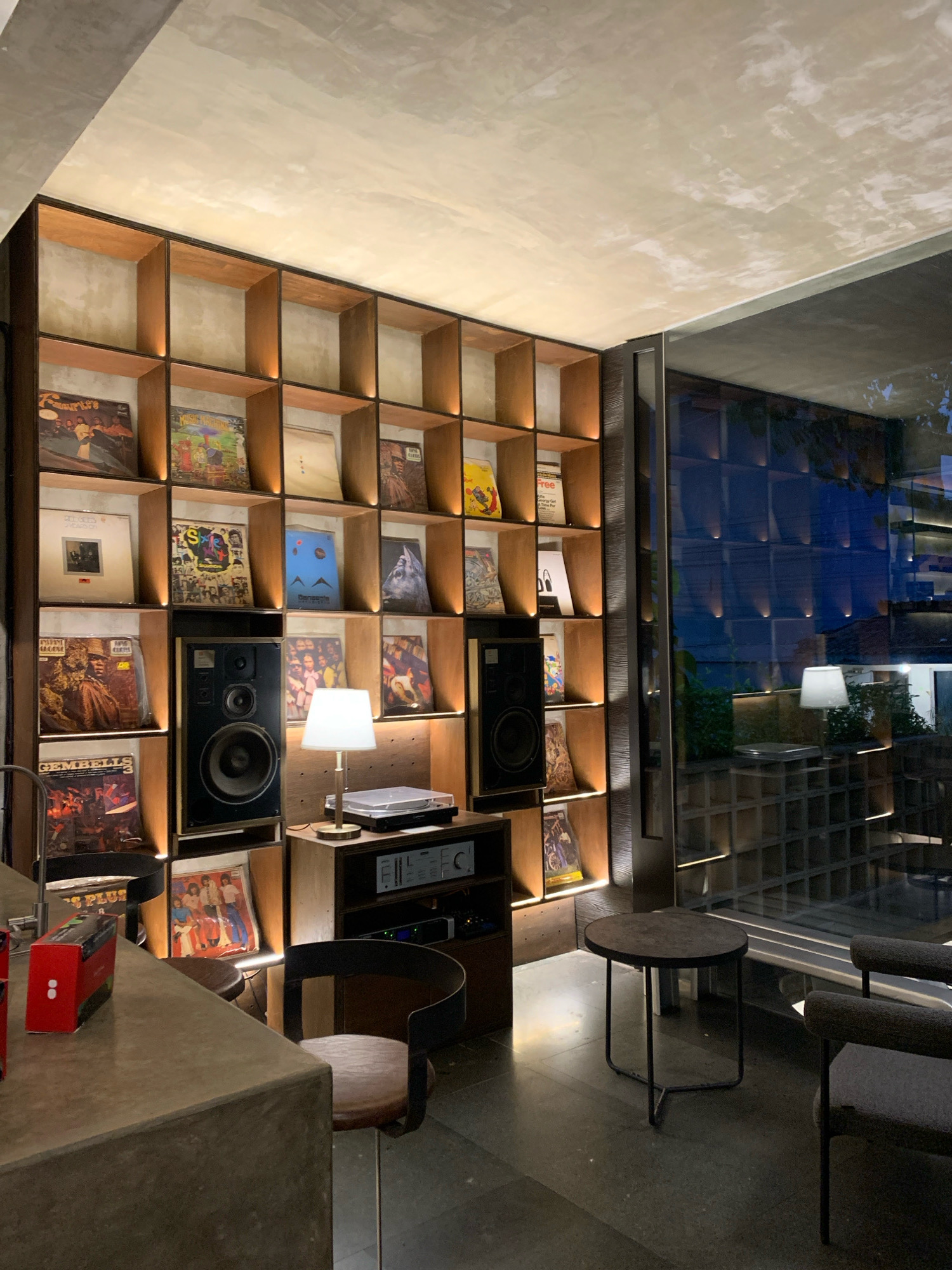 The image shows a well-lit, modern room with a large bookshelf filled with vinyl records. The bookshelf is made of wood with individual cubbies for each record. Some of the records have colorful album art visible, while others are obscured. Two large black speakers are placed on shelves within the bookshelf. In front of the bookshelf, there is a wooden cabinet with audio equipment, including a turntable and an amplifier. A small lamp with a white shade sits on top of the cabinet. To the right of the bookshelf, a large window offers a view of another room with a grid-like pattern on the wall and subtle lighting. In the foreground, a concrete counter is visible, with a red box and a smaller box on it. A wooden chair with a dark seat is positioned near the counter. A small, dark, round table with a metal base stands on the floor next to a dark-colored armchair. The floor appears to be dark concrete. The room has a minimalist aesthetic with a focus on natural materials and clean lines. There is no indication of the city of Mojokerto, Indonesia in the image.