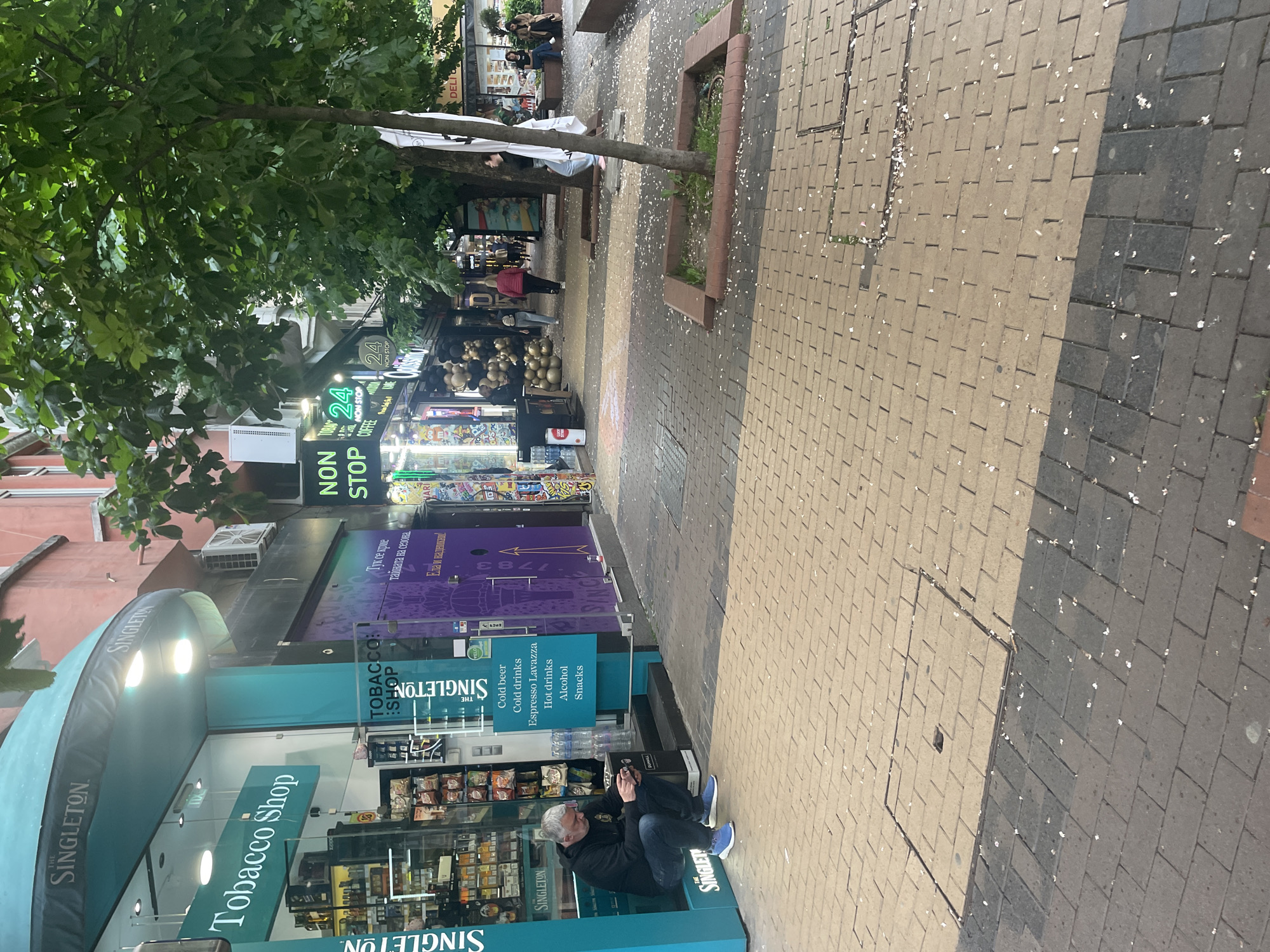 The image shows a street scene in Sofia, Bulgaria. A man is sitting on the sidewalk near a "Singleton Tobacco Shop" and next to a "24 Non Stop" store. The street is paved with brick, and there are trees and other shops in the background. Several persons are walking in the background. The storefronts have signs written in Bulgarian and English, indicating that cold beer, cold drinks, espresso Lavazza, hot drinks, alcohol, and snacks are available.