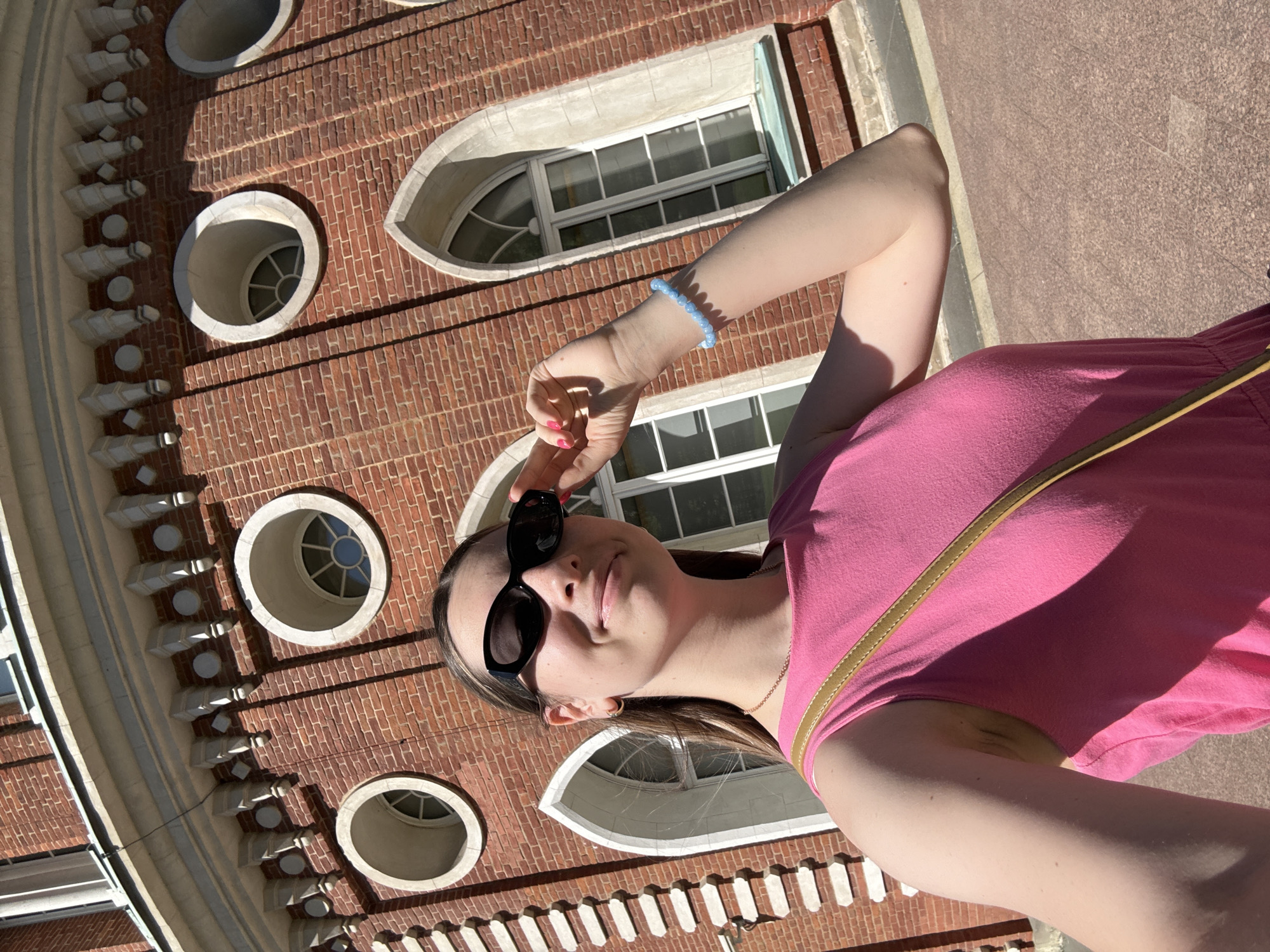 A young woman with dark hair, wearing sunglasses, a pink tank top, and a gold crossbody bag, stands in front of a red brick building with arched windows and circular openings. She is holding her sunglasses with her right hand, and a blue beaded bracelet is visible on her left wrist. The building appears to be a historical structure, possibly a palace or estate, with ornate architectural details. The ground in front of the building is paved with reddish-brown tiles. The image is taken outdoors on a sunny day. This architectural style is characteristic of Tsaritsyno Park in Moscow, Russia.