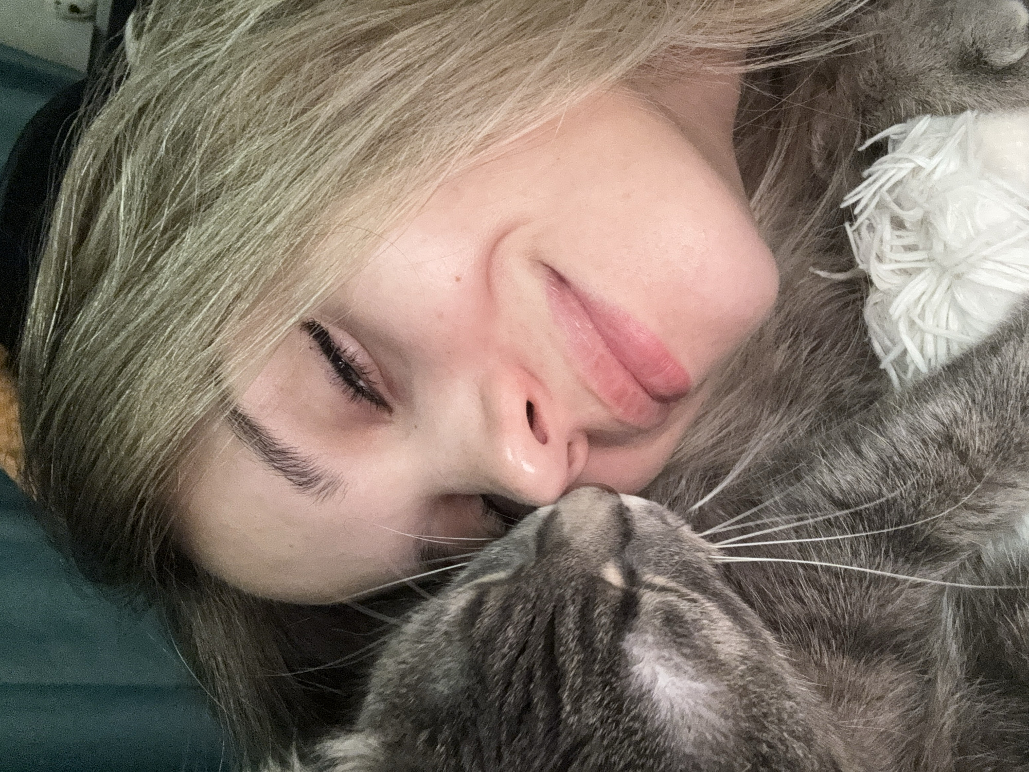 A close-up shows a woman with fair skin and light blonde hair lying down, her eyes mostly closed with dark eyelashes visible. She has a slight smile, and her lips are pink. Her nose is close to the head of a gray tabby cat. The cat is facing towards her, its whiskers prominent. The background appears to be a dark green or teal surface. The location is not specified.