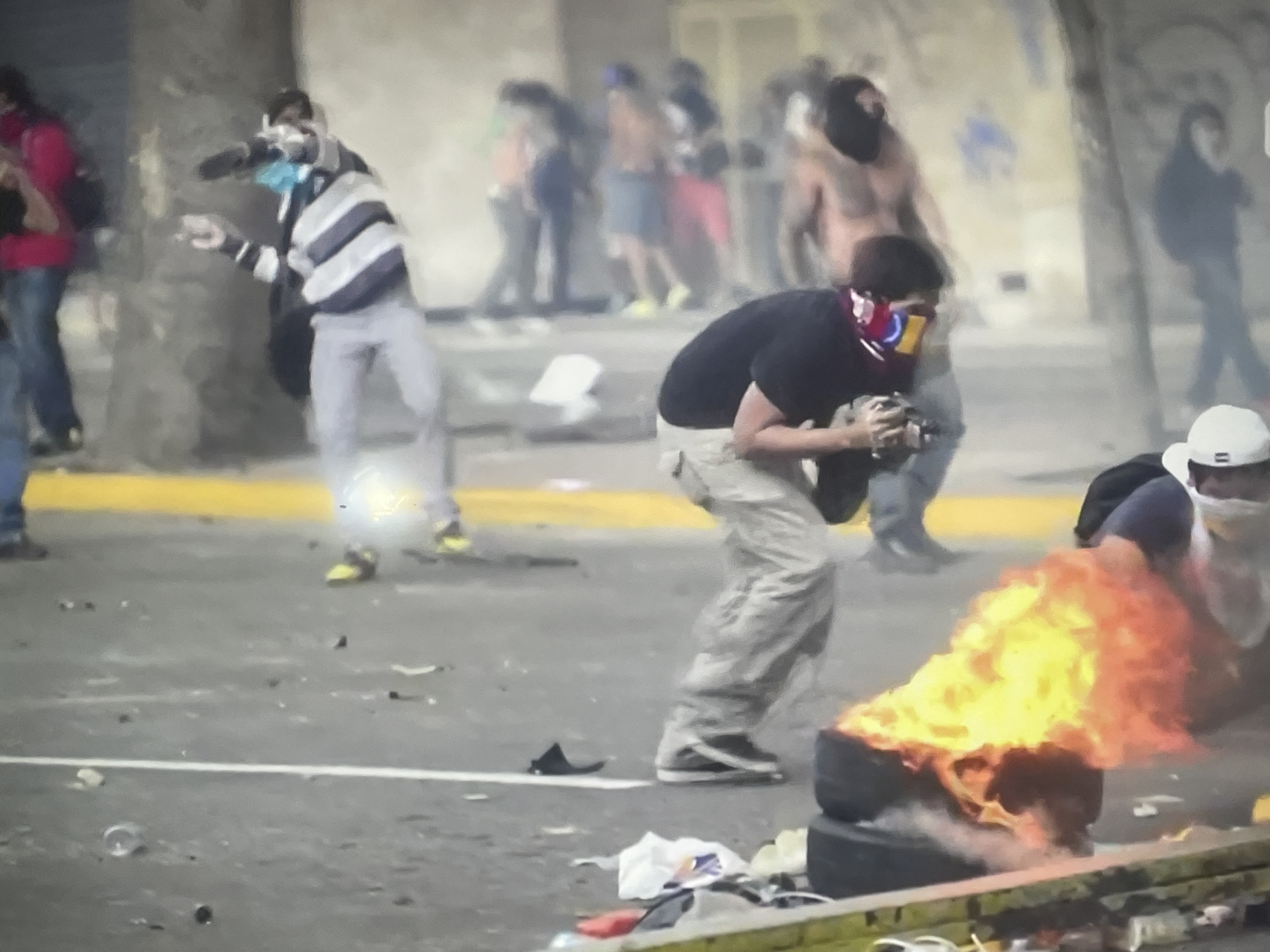 The image depicts a street scene in Unknown, Unknown, appearing to show a protest or riot. Several people are present, some wearing masks or scarves covering their faces. One individual is positioned near a burning tire, while others are seen in the background. One person appears to be throwing an object. The environment shows signs of unrest with debris scattered on the ground. The overall scene conveys a sense of chaos and conflict.