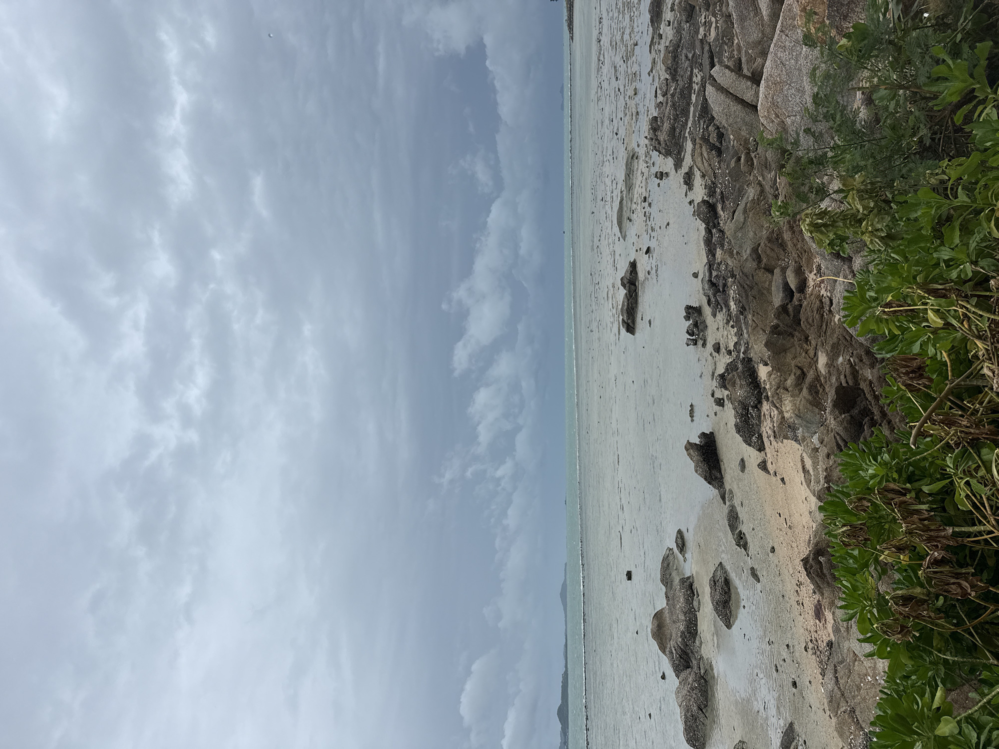 An eye-level, medium shot captures a rocky beach on the coast of Ko Samui District, Thailand under a cloudy sky. The foreground is dominated by large, grey rocks and vibrant green foliage. The middle ground shows a sandy, wet beach dotted with smaller rocks and shallow water. In the background, the sea stretches out to a distant, hazy shoreline with hills. The sky is filled with heavy, grey clouds, suggesting an overcast or approaching storm. There are no people or animals visible in the image.