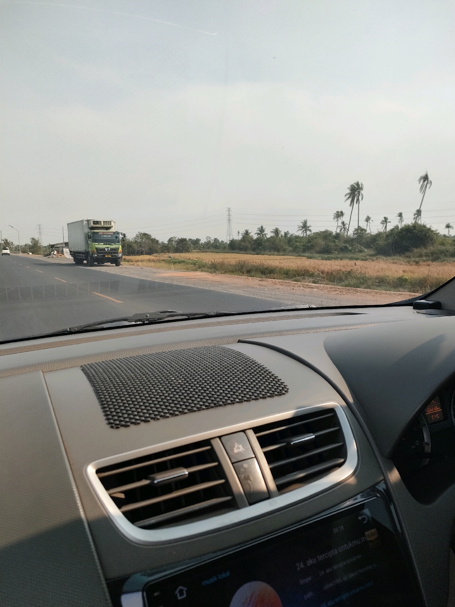 The image is taken from the interior of a vehicle looking out at a highway scene in Lok Baintan Dalam, Indonesia. In the foreground, the dashboard of the car is visible, including air vents and a digital display showing the time as 08:18. A large green truck with a white refrigerated trailer is traveling on the right lane of the highway. The highway is flanked by dry, brown vegetation and scattered palm trees under a bright, hazy sky. Power lines and transmission towers are visible in the distance. The road appears to be in a rural or semi-rural area.