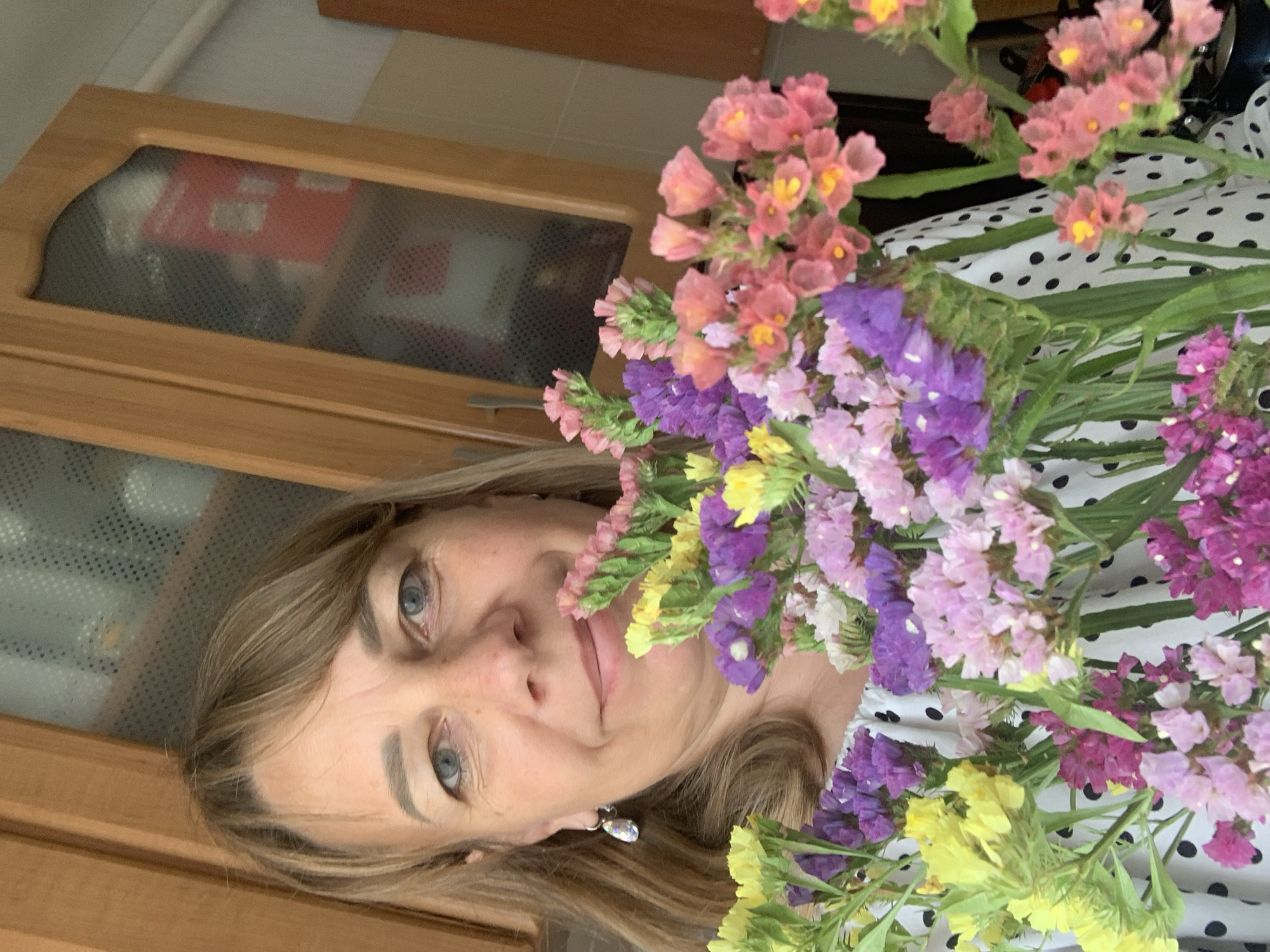 A blonde woman with blue eyes and fair skin is holding a bouquet of dried flowers in front of her chest. She is wearing a white top with black polka dots and a diamond-shaped earring in her left ear. The background consists of wooden cabinets with glass doors and a light-colored wall. The scene appears to be indoors, likely in a kitchen or living room, given the cabinetry. The image quality is moderate, with some blurriness and graininess. The woman is looking directly at the camera and smiling slightly. The bouquet is composed of flowers in various shades of pink, purple, and yellow. The lighting is natural and appears to be coming from the front. There is no specific indication of the location within Kapyĺ, Belarus, based solely on the visual information provided in the image.
