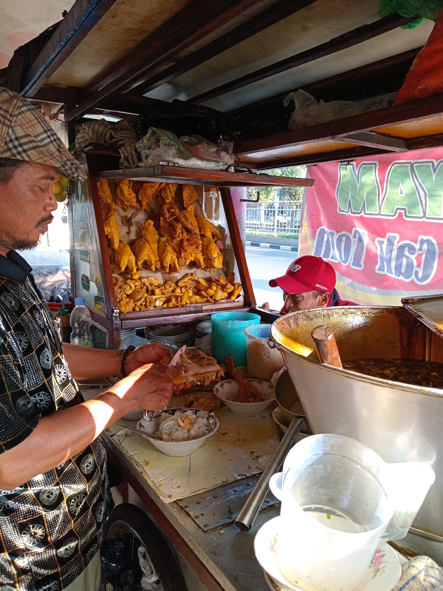 A man wearing a plaid flat cap and a patterned shirt is preparing food at a street food stall in Kroman, Indonesia. He is slicing meat, likely chicken, with a knife. Behind him, whole fried chickens are displayed. Another man, wearing a red baseball cap, is visible in the background, looking towards the food. The stall appears to be on a street, with a red banner displaying text in green and white behind the men. Various containers, bowls, and a large pot are present on the counter. The scene suggests a typical outdoor food vending operation.