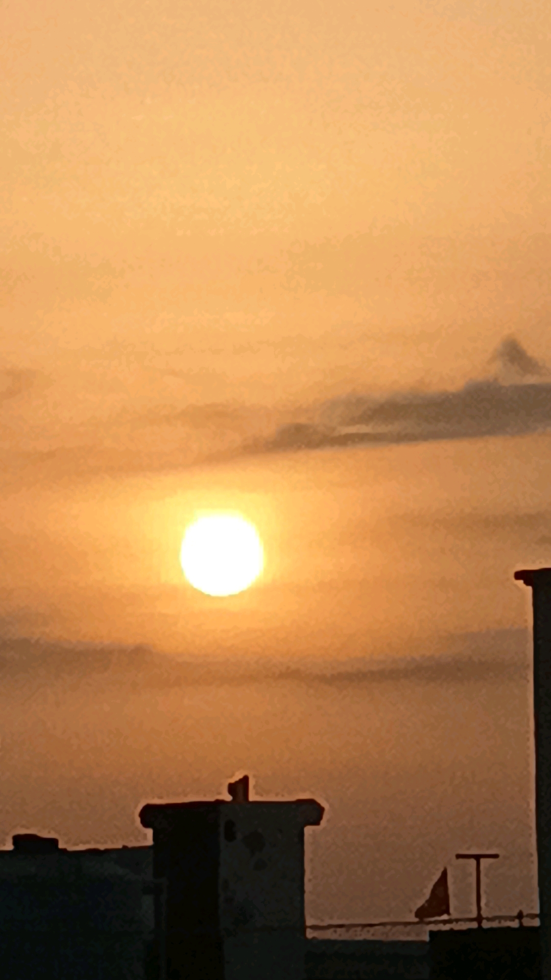 This image captures a sunset over a city skyline in Unknown, Unknown. The sun, a bright orb, is prominently displayed in the center of the frame, casting a warm, orange glow across the sky. Wispy clouds are scattered throughout, some dark and appearing to be on the verge of dissipating. In the foreground, the silhouetted rooftops of buildings are visible, their dark shapes contrasting with the vibrant sky. One building in particular features a chimney-like structure with a distinct, almost angular top. Another structure further to the right displays a small, triangular flag and a simple, T-shaped antenna. The overall impression is one of a peaceful evening in an urban setting.