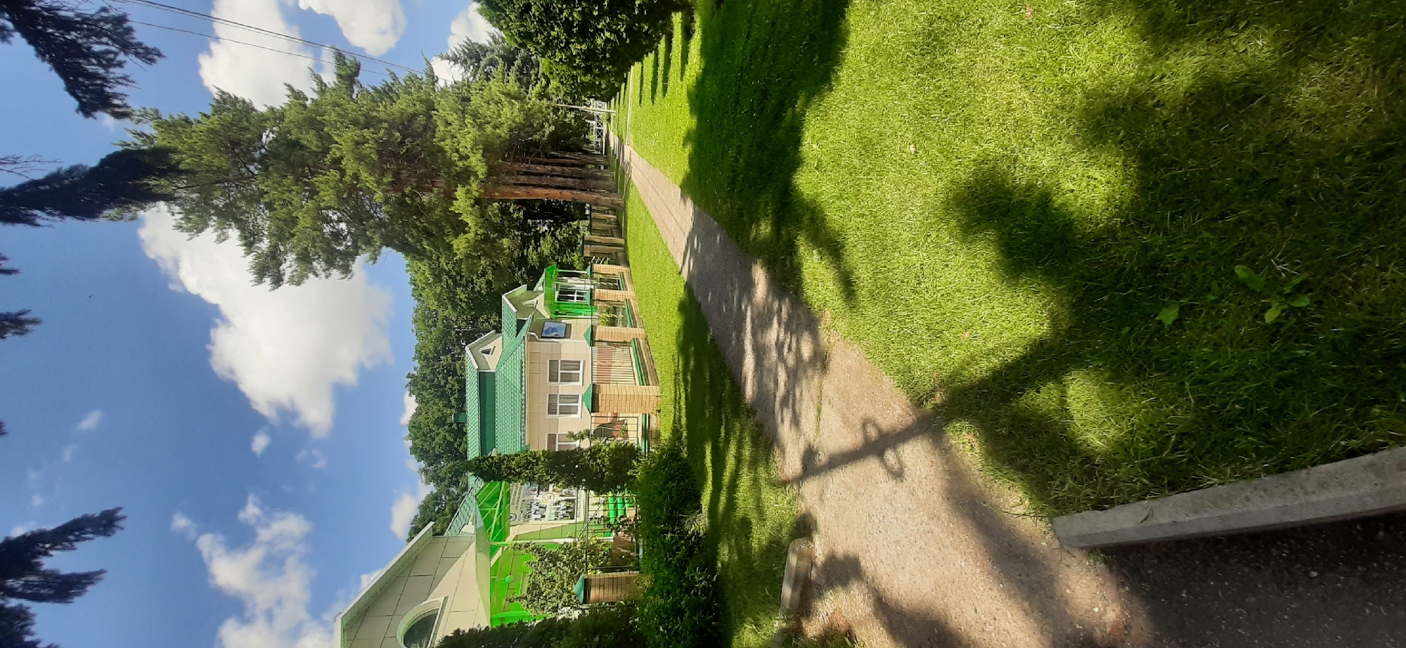The image depicts a sunlit pathway running alongside a neatly manicured lawn and a low fence. Beyond the fence, there are buildings with green roofs and light-colored walls, surrounded by lush green foliage and tall trees. The pathway is made of gravel and is partially shaded by tree branches casting shadows. The sky above is a vibrant blue with scattered white clouds. The location is identified as Yessentukskaya, Russia. There are no people visible in the image.