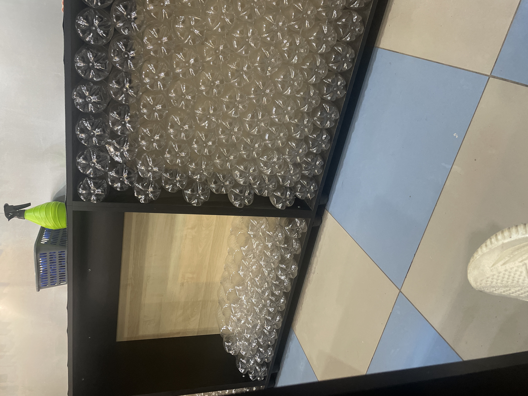 The image shows a shelf unit filled with clear plastic bottles. The shelf unit is black and has two sections. The left section is empty except for a wooden back panel. The right section is filled with rows of plastic bottles, stacked in a diamond pattern, facing upwards. On top of the shelf unit, to the left, is a gray plastic basket and a green spray bottle. The floor is tiled in a pattern of beige and blue squares. The scene is located in Zhukovo, Russia.