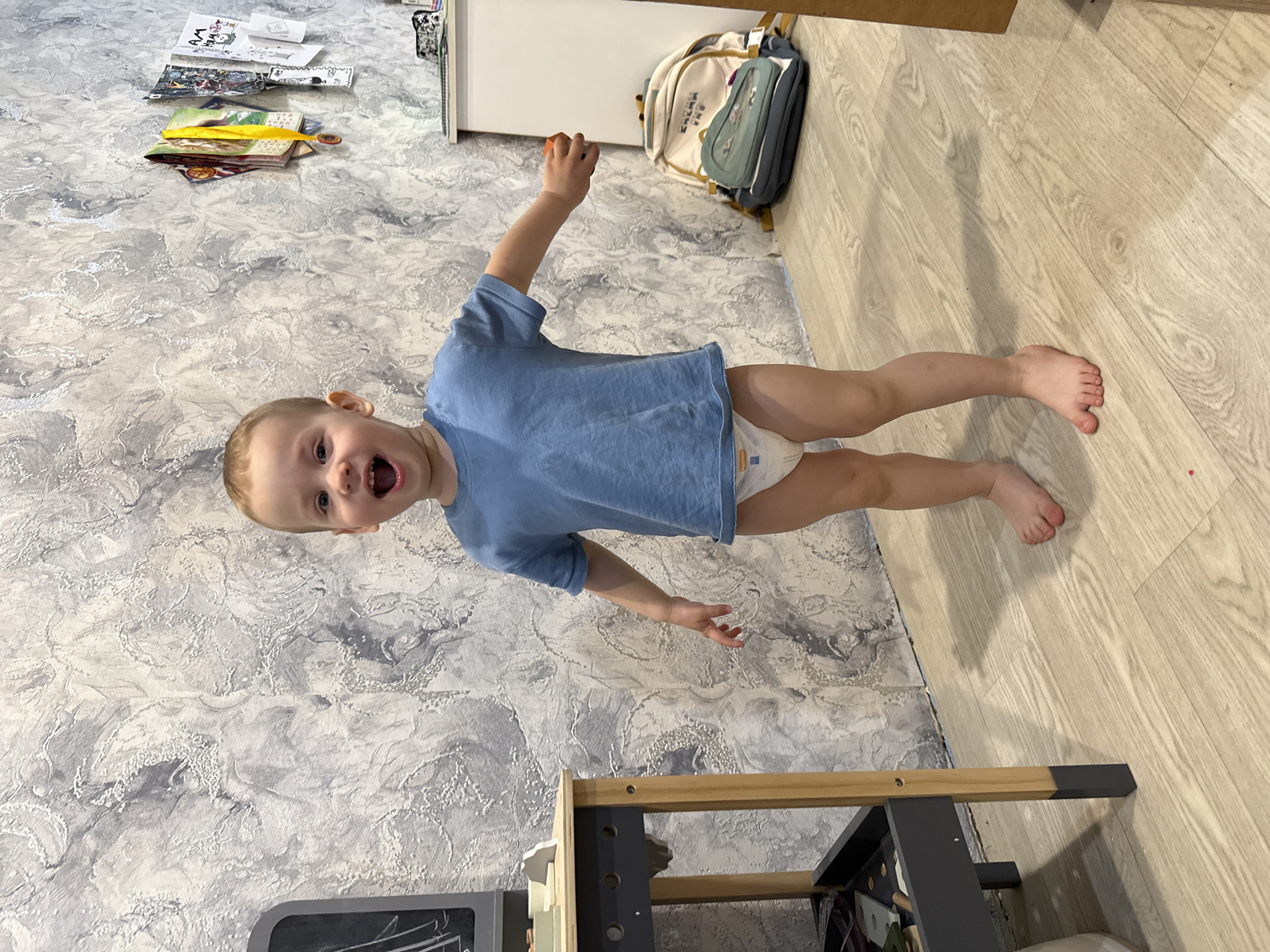 A young child stands on a floor that is a mix of gray-patterned wallpaper-like material and light brown wood flooring. The child wears a blue t-shirt and a diaper. The child has short blond hair and is smiling with their mouth open. The child's left arm is raised.
A backpack, books, and papers are on the floor. A light brown wooden structure with dark gray legs is in the foreground. Rusanovo, Russia is not mentioned.