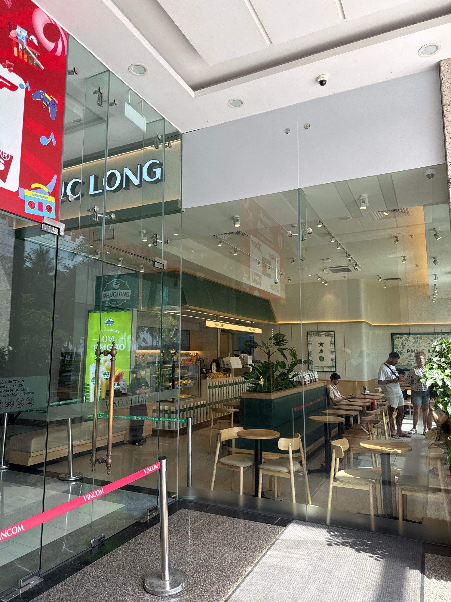 The image shows the entrance to a Phúc Long coffee shop located in Nha Trang, Vietnam, a city on Vietnam's central coast. The entrance is made of glass doors and windows, with a red velvet rope barrier in front. Inside, patrons can be seen seated at tables, some ordering, and others enjoying their drinks. The interior decor features green accents, wooden furniture, and plants, creating a modern and inviting atmosphere. The Phúc Long logo is prominently displayed on signage and on cups. The overall scene suggests a bustling cafe in a city known for its beaches and tourism.
