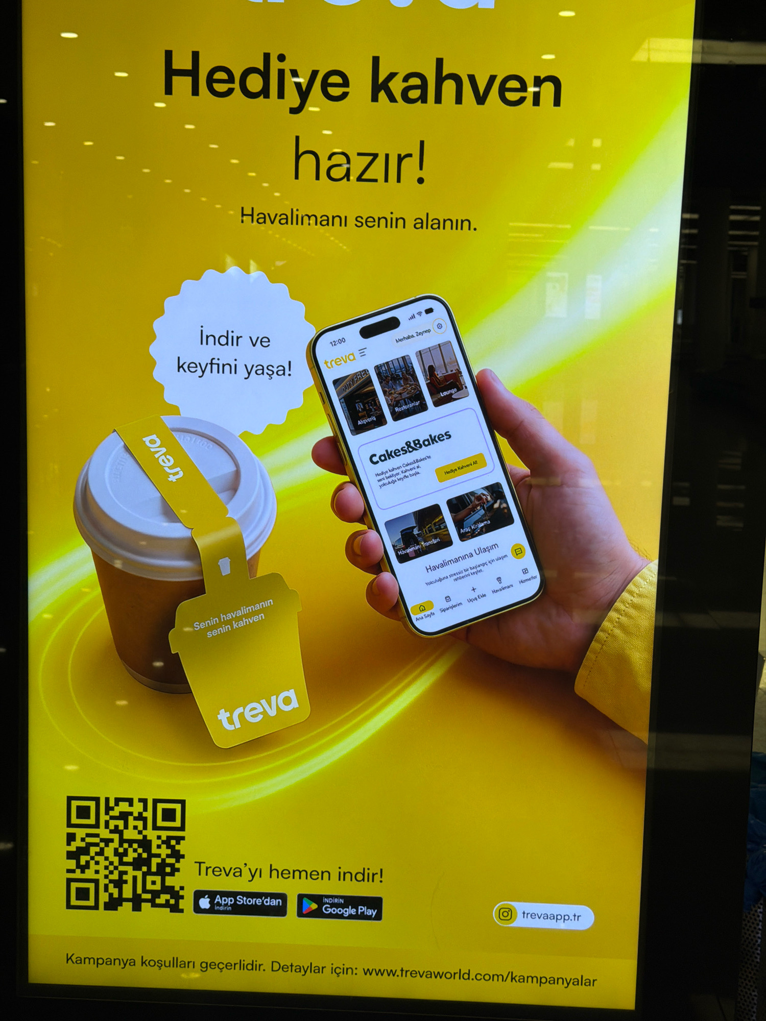 The image is an advertisement displayed on a large, illuminated screen, likely in a public space. The dominant color is bright yellow, with black text. The advertisement promotes a mobile application called "Treva," which offers services related to airports and travel.
At the top, in large black lettering, it says "Hediye kahven hazır!" which translates to "Your gift coffee is ready!" Below that, "Havalimanı senin alanın." means "The airport is your domain."
The central part of the advertisement features a hand holding a smartphone, displaying the Treva app interface. The app shows various categories like "Restoranlar" (Restaurants), "Lounge," "Cakes&Bakes," and "Araç Kiralama" (Car Rental). A prominent section on the app is titled "Havalimanına Ulaşım" (Airport Transportation), offering to make travel stress-free.
To the left of the phone is a takeaway coffee cup with a yellow tag that says "treva" and has the tagline "Senin havalimanın senin kahven" (Your airport, your coffee).
At the bottom of the screen, there is a QR code, and text encouraging users to "Treva'yı hemen indir!" (Download Treva now!). Below this are icons for the App Store and Google Play, with text indicating download options. The website address "www.trevaworld.com/kampanyalar" is also provided. Additionally, an Instagram handle "@trevaapp.tr" is visible.
The overall scene is a digital advertisement designed to attract users to download and utilize the Treva app for airport-related services. The mention of "Gaziemir, Turkey" in the context of this image is not supported by any visual cues within the advertisement itself. The advertisement is a general promotion.