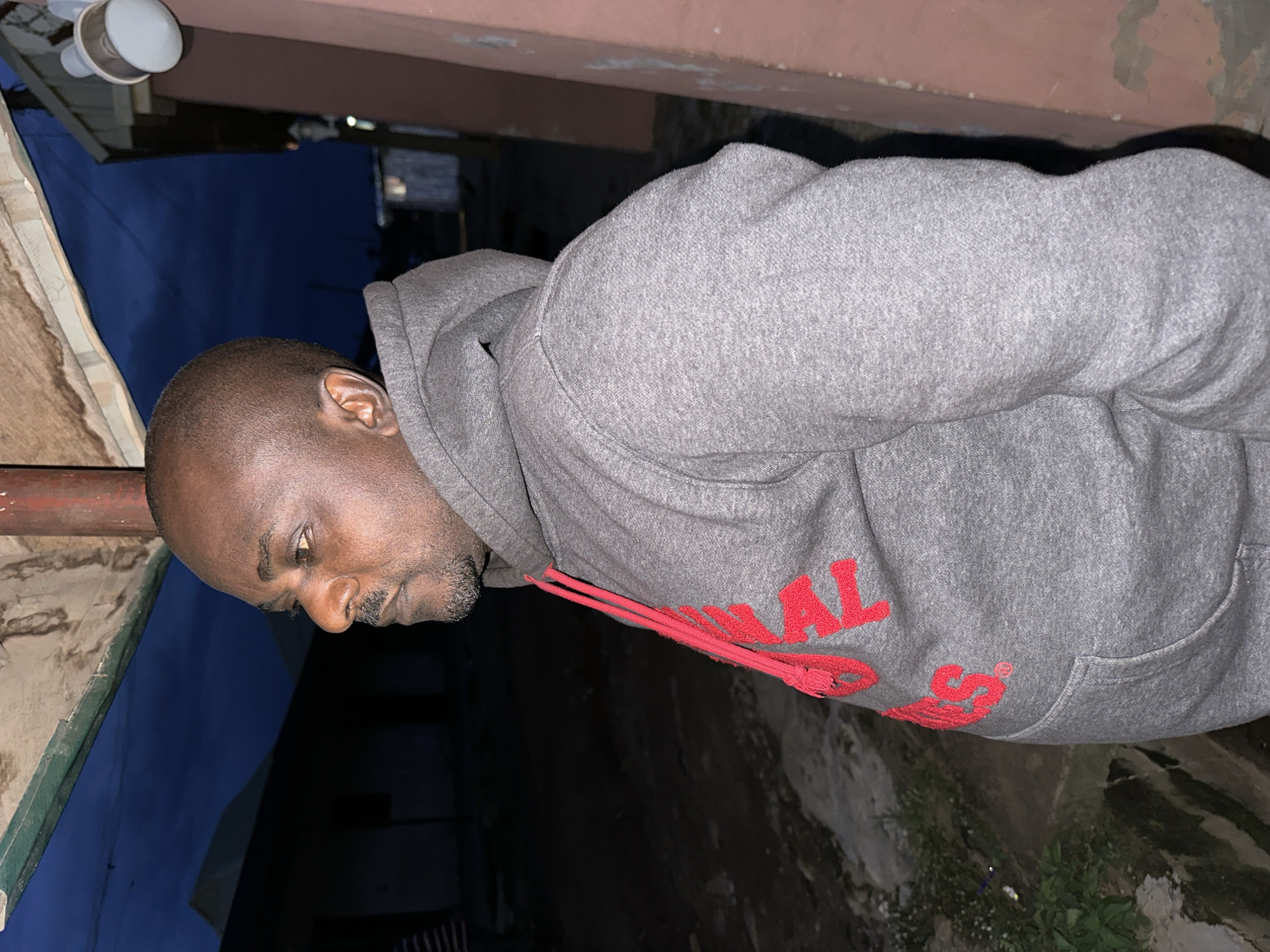 An adult male is captured in a medium shot, positioned on the right side of the frame, looking to the left. He has dark skin, a shaved head, and is wearing a grey hooded sweatshirt with red lettering on the front and red drawstrings. He has a goatee and mustache. The background suggests an outdoor setting at dusk or night, with a dark blue sky visible above. Structures, possibly buildings, are dimly lit in the background. The foreground shows a dirt or gravel ground with some sparse green vegetation. The overall environment appears to be a somewhat neglected outdoor area, potentially on the outskirts of a settlement. This scene is likely located in the Narayi, Nigeria area.