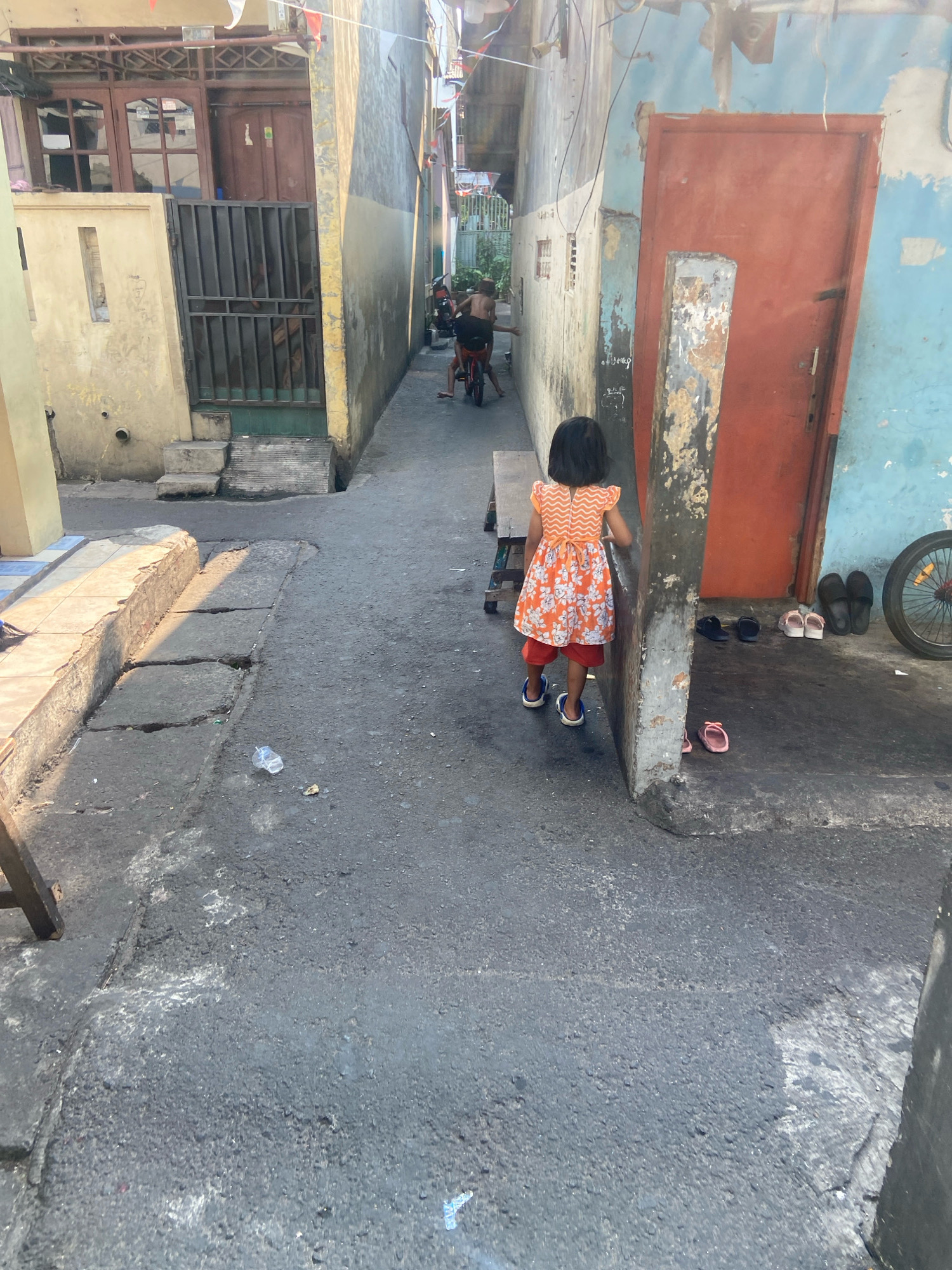 The image depicts a narrow alleyway in a densely populated urban area, likely a residential neighborhood in North Jakarta, Indonesia. The buildings lining the alley are close together, with weathered facades suggesting age and exposure to the elements. One building on the right features a large, weathered orange door and a section of pale blue painted wall. Adjacent to this, a concrete pillar stands prominently. Several pairs of sandals and flip-flops are arranged on the ground near the door, indicating it is an entrance to a dwelling.
In the foreground, a young girl wearing an orange and white floral dress over red shorts and blue sandals walks away from the camera, her back to the viewer. She appears to be walking towards the deeper part of the alley. To the left, a damaged concrete ledge runs along the side of the alley, with cracks and discolored patches. A discarded plastic bottle lies on the asphalt surface.
Further down the alley, another child, partially visible from the back and wearing dark shorts, is riding a red bicycle. The alley floor is paved with dark asphalt, showing signs of wear and tear, including cracks and uneven surfaces. The overall scene conveys a sense of everyday life in a working-class urban environment, characterized by close living quarters and the presence of children engaged in outdoor activities. The lighting suggests it is daytime, with sunlight casting shadows on the alley floor.