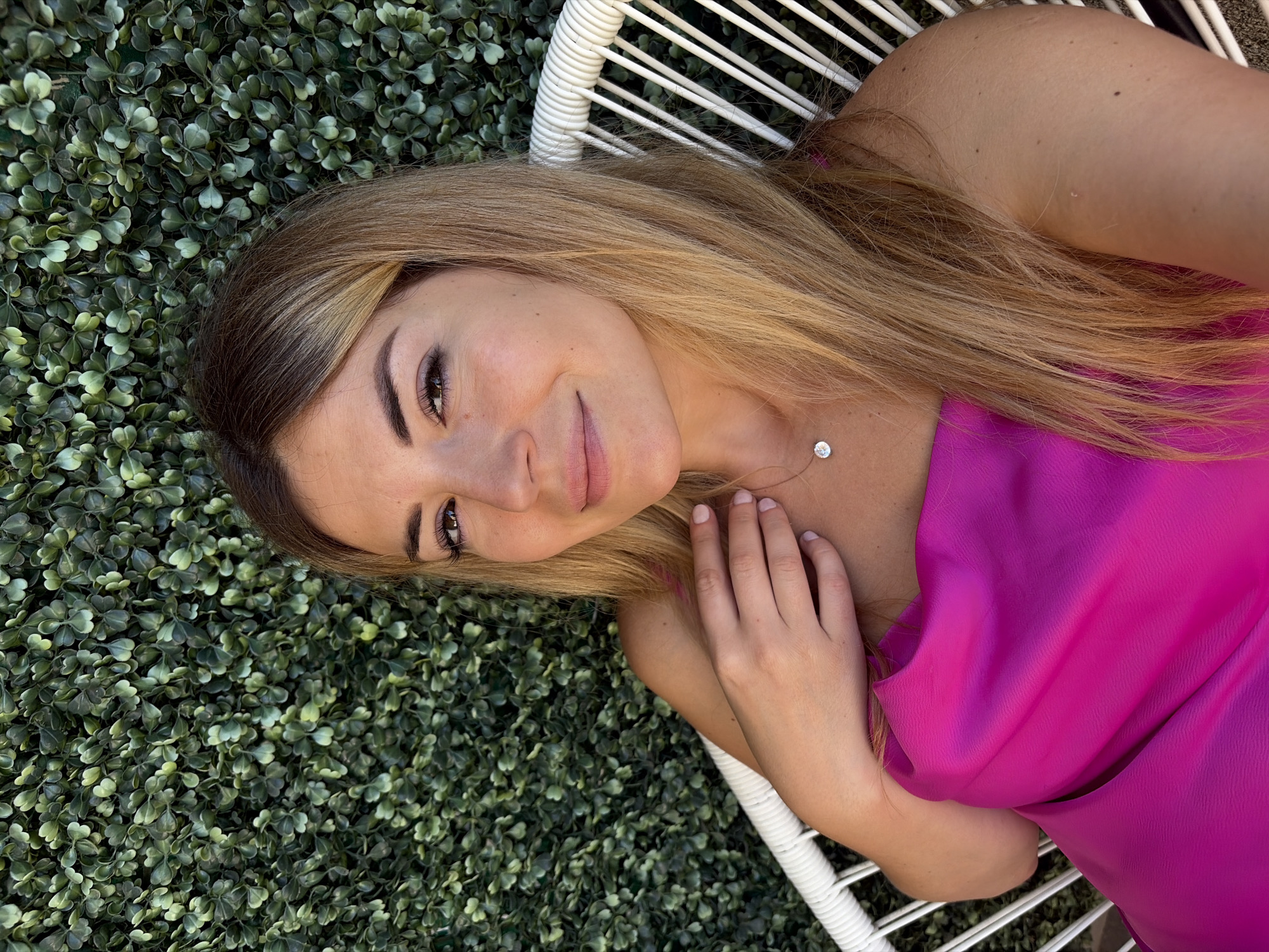 A young adult female with blonde hair and light skin is seated on a white wicker chair, facing forward and slightly to her left. She is wearing a bright pink dress and a delicate silver necklace with a single diamond pendant. Her right hand is resting on her chest. Behind her is a dense green hedge wall, characteristic of outdoor decorative elements found in many urban settings, including upscale establishments in the city of Budva, Montenegro. The lighting suggests it is daytime, with natural light illuminating her face and the scene.