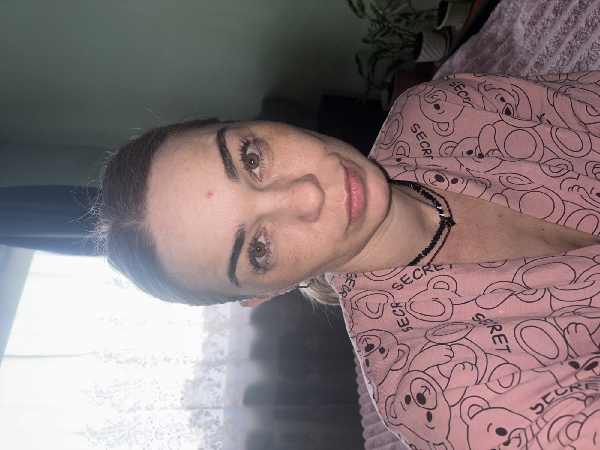 A woman with light skin, brown hair pulled back, and green eyes is captured in a medium close-up shot, looking directly at the camera. She has a light smattering of freckles on her face and a small red mark on her forehead. Her lips are slightly parted. She is wearing a pink pajama top adorned with a pattern of black teddy bear outlines and the word "SECR" repeated. A dark beaded necklace is visible around her neck. The background is softly blurred, revealing a green wall on the right, and on the left, a window with sheer white curtains. Potted plants are visible in the background on the right. The scene suggests an indoor, possibly domestic, setting.